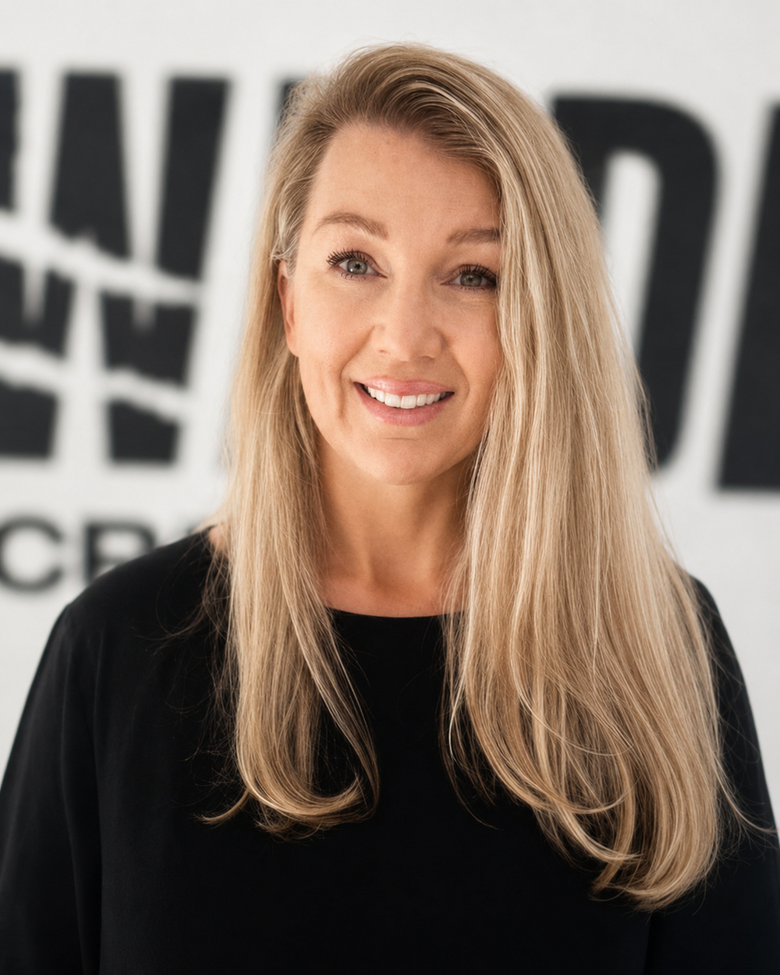 A woman with long blonde hair smiling, wearing a black top, standing in front of a blurred background with partially visible black and white text  in front of a blurred logo that says 'Wildlings'