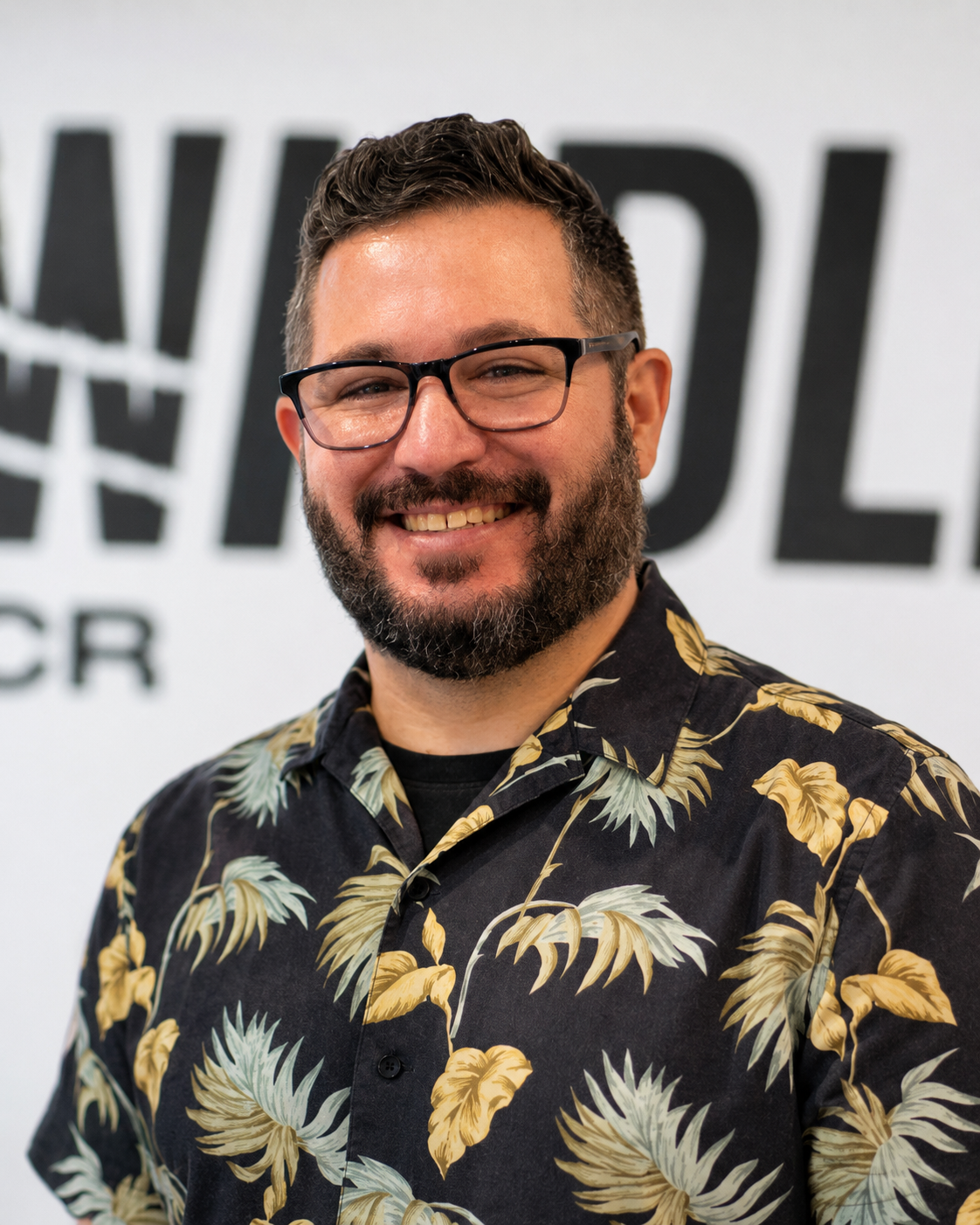 A smiling man with glasses and a beard wearing a black Hawaiian shirt with a floral pattern, standing in front of a white background with black text.
