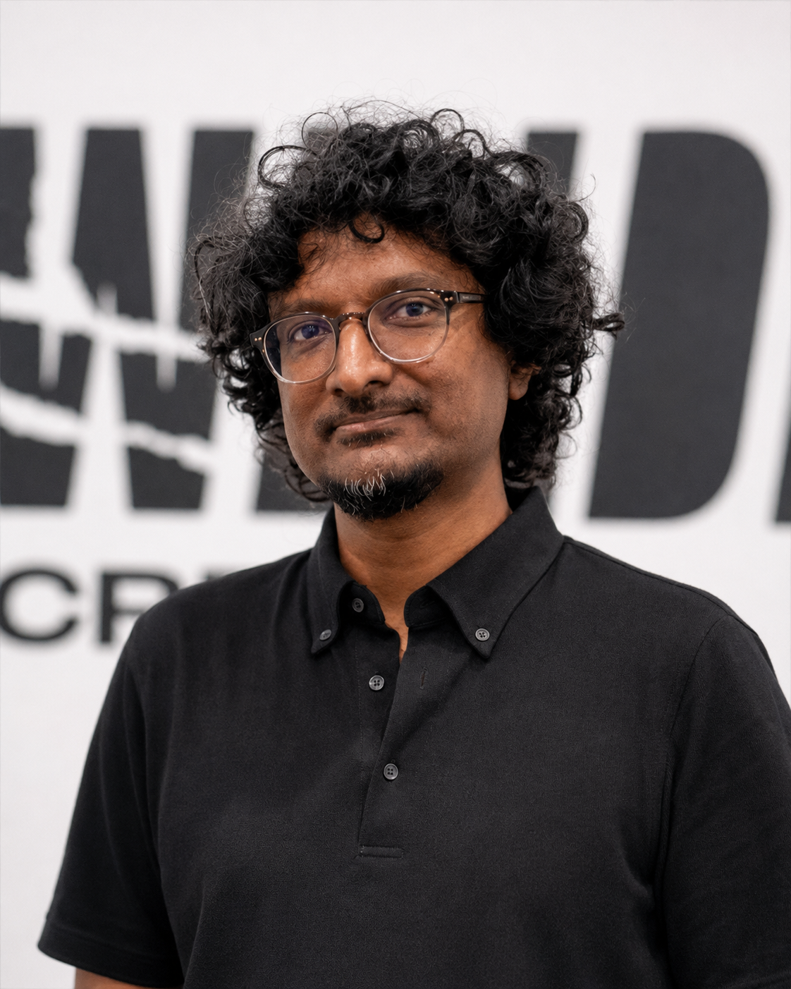 A man with curly black hair, wearing glasses and a black collared shirt, standing in front of a wall with black and white text in the background.