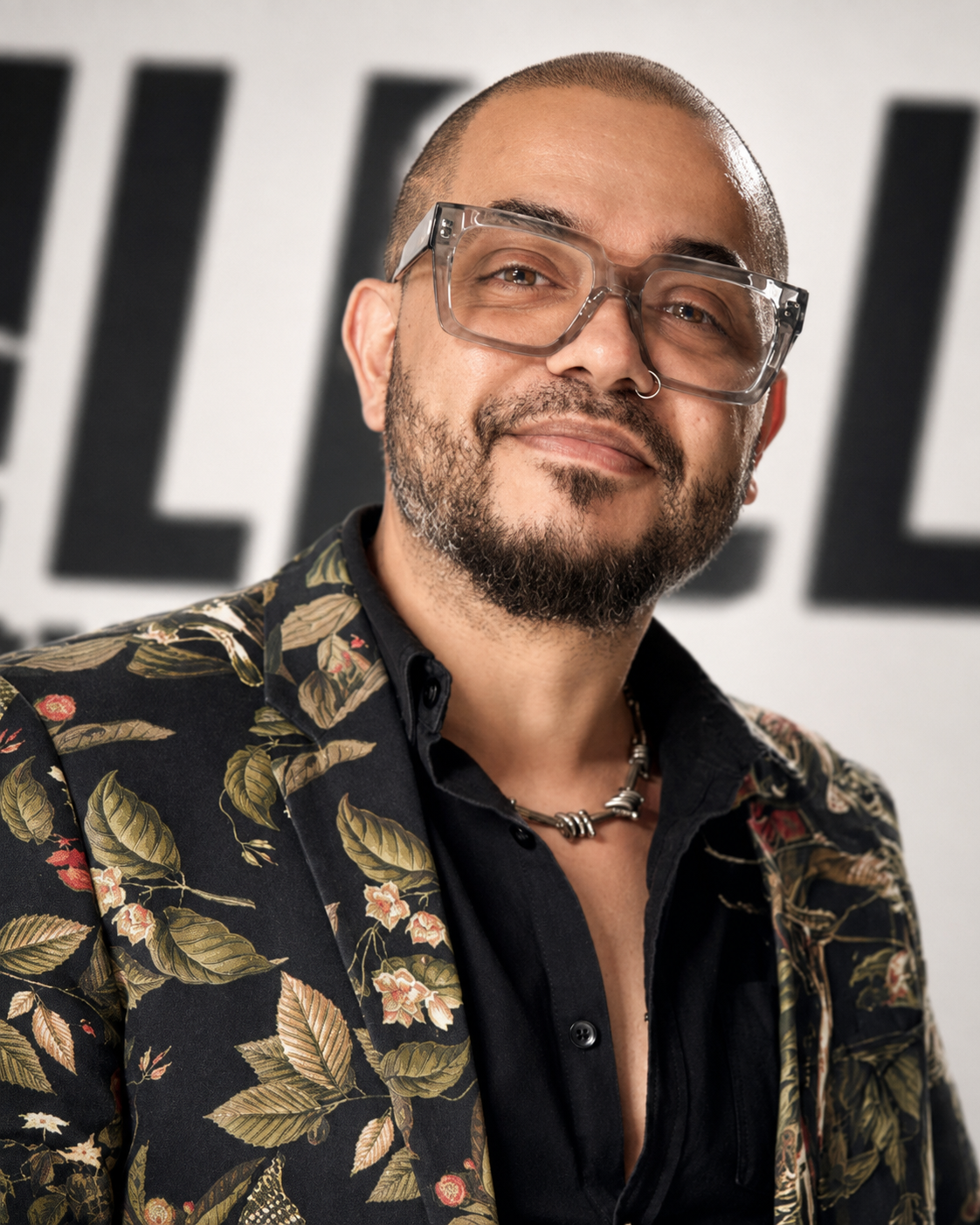 A man with a beard and glasses, wearing a floral blazer and a beaded necklace, standing in front of a partially visible sign.