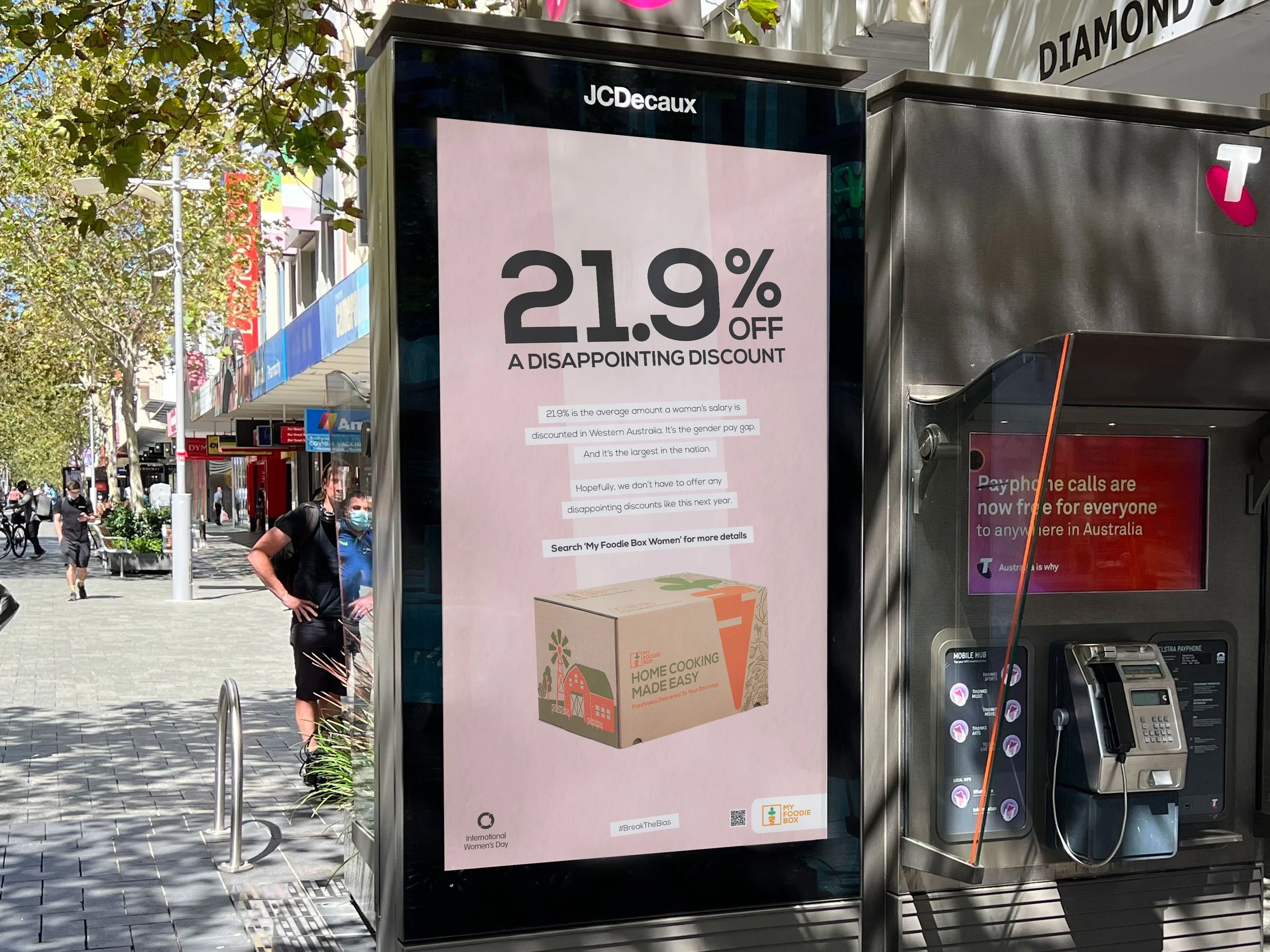 A street scene with a digital sign advertising a 21.9% discount, cars, pedestrians, and storefronts in the background, including an advertisement for home cooking boxes.