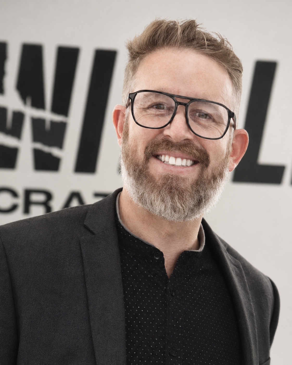 A smiling man with glasses, a beard, and short hair wearing a black blazer and black shirt, standing in front of a blurred background with large black text.