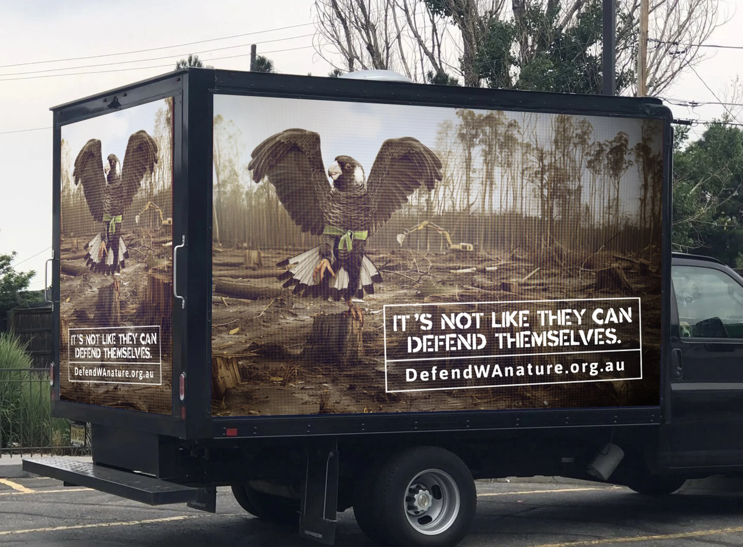 Black truck with a large poster on each side depicting a bird with open wings, standing on a landscape of fallen logs and trees. Text on the posters states, 'It's not like they can defend themselves.' and the website URL defendwanature.org.au.