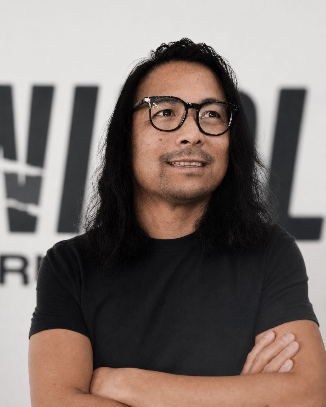 A man with long black hair, wearing glasses and a black t-shirt, standing with arms crossed in front of a background with large black text.