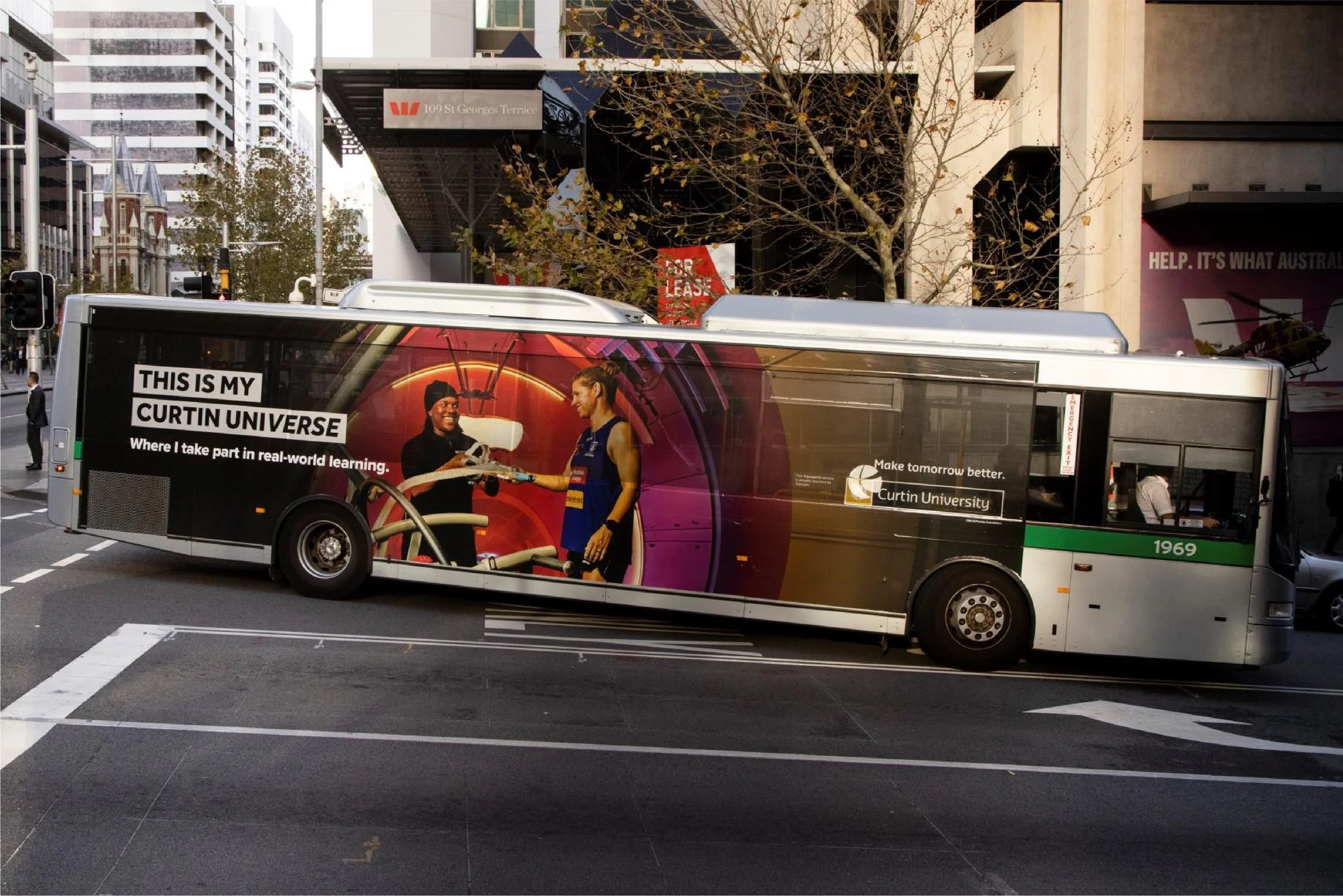 A city bus with an advertisement for Curtin University, featuring two smiling women, one wearing a cap and the other in athletic clothes, inside an urban environment with tall buildings and trees.