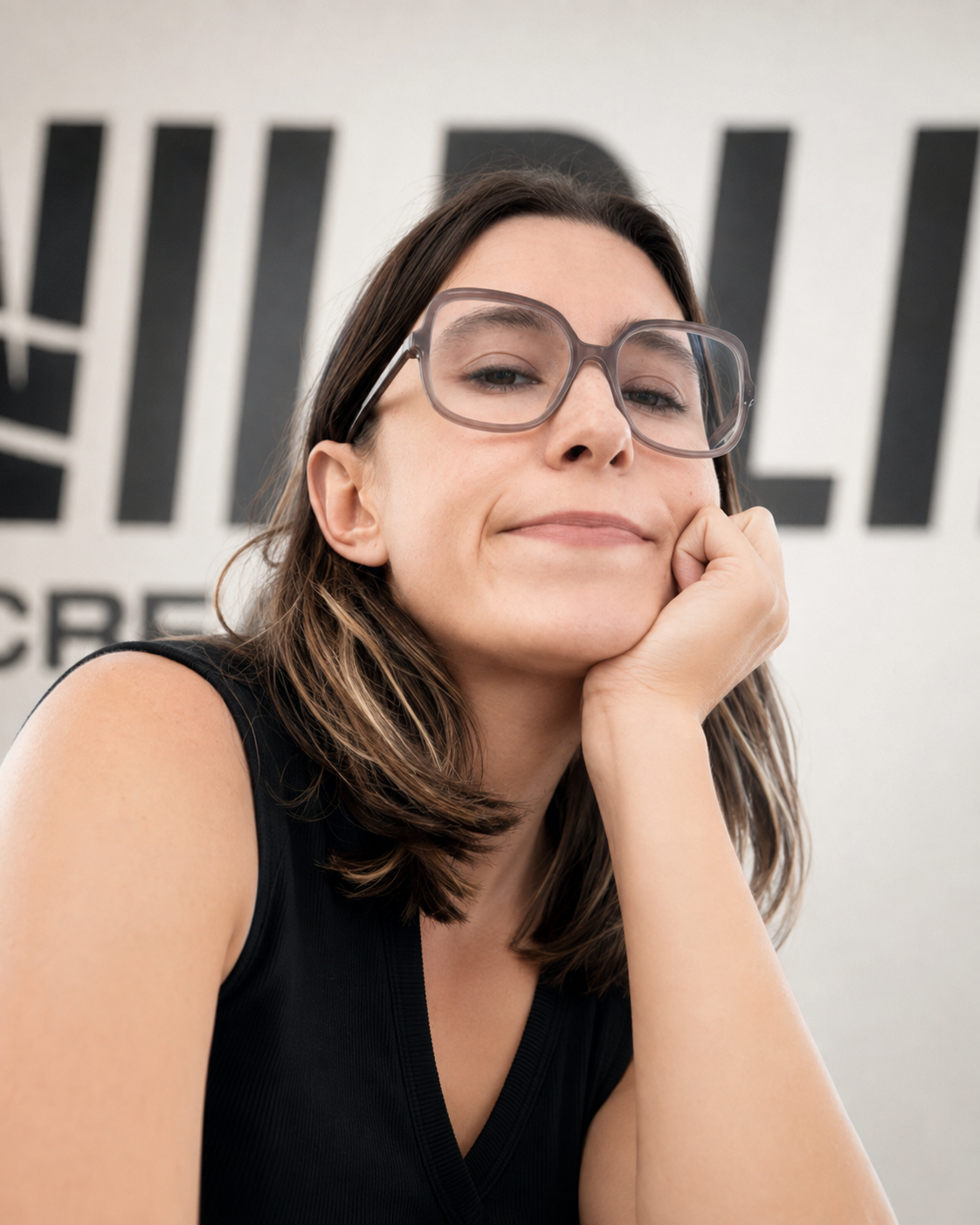 Close-up of a woman with shoulder-length brown hair, wearing glasses, resting her chin on her hand, with a neutral expression and a large black and white sign in the background.