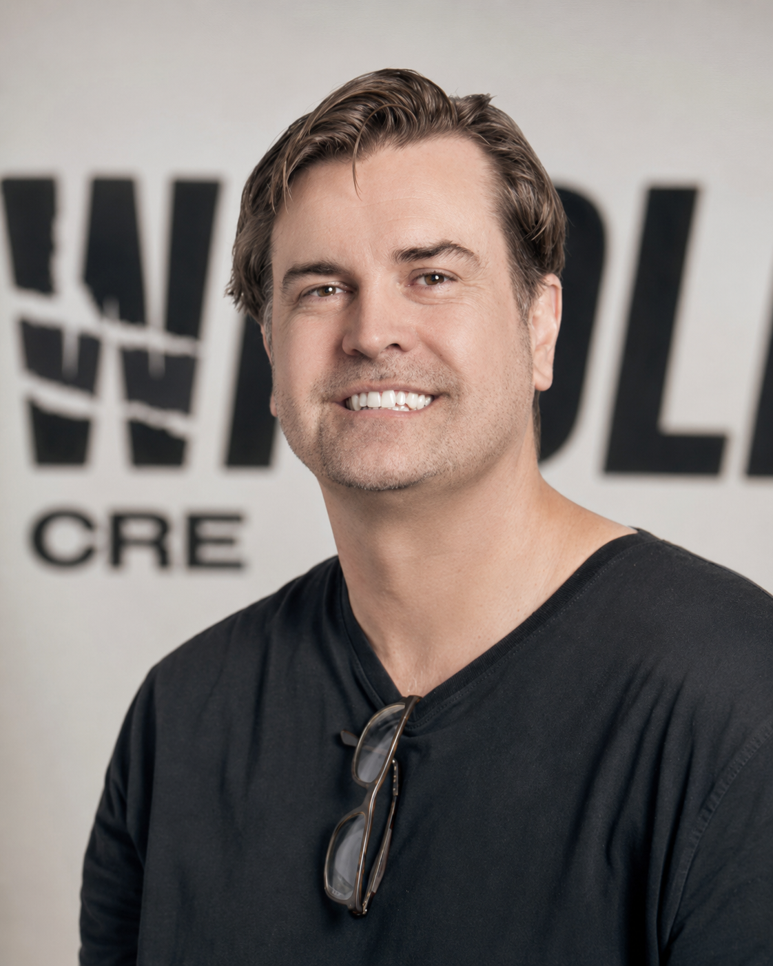 A smiling man with brown hair and glasses hanging from his shirt, wearing a black shirt, standing in front of a wall with the words 'WALL' and 'CRE' partially visible.