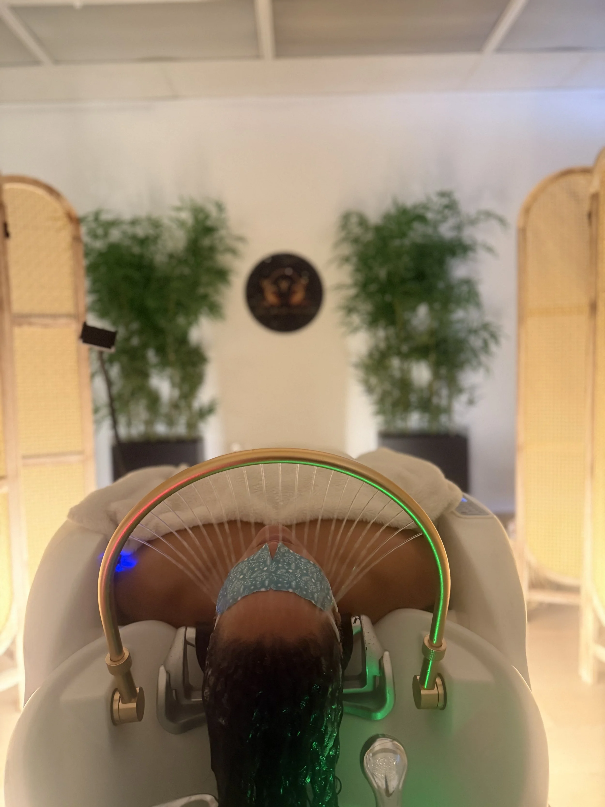 A person lying in a medical imaging machine with a circular opening, with glowing green and blue lights around the machine, and a wall with plants and a decorative piece in the background.