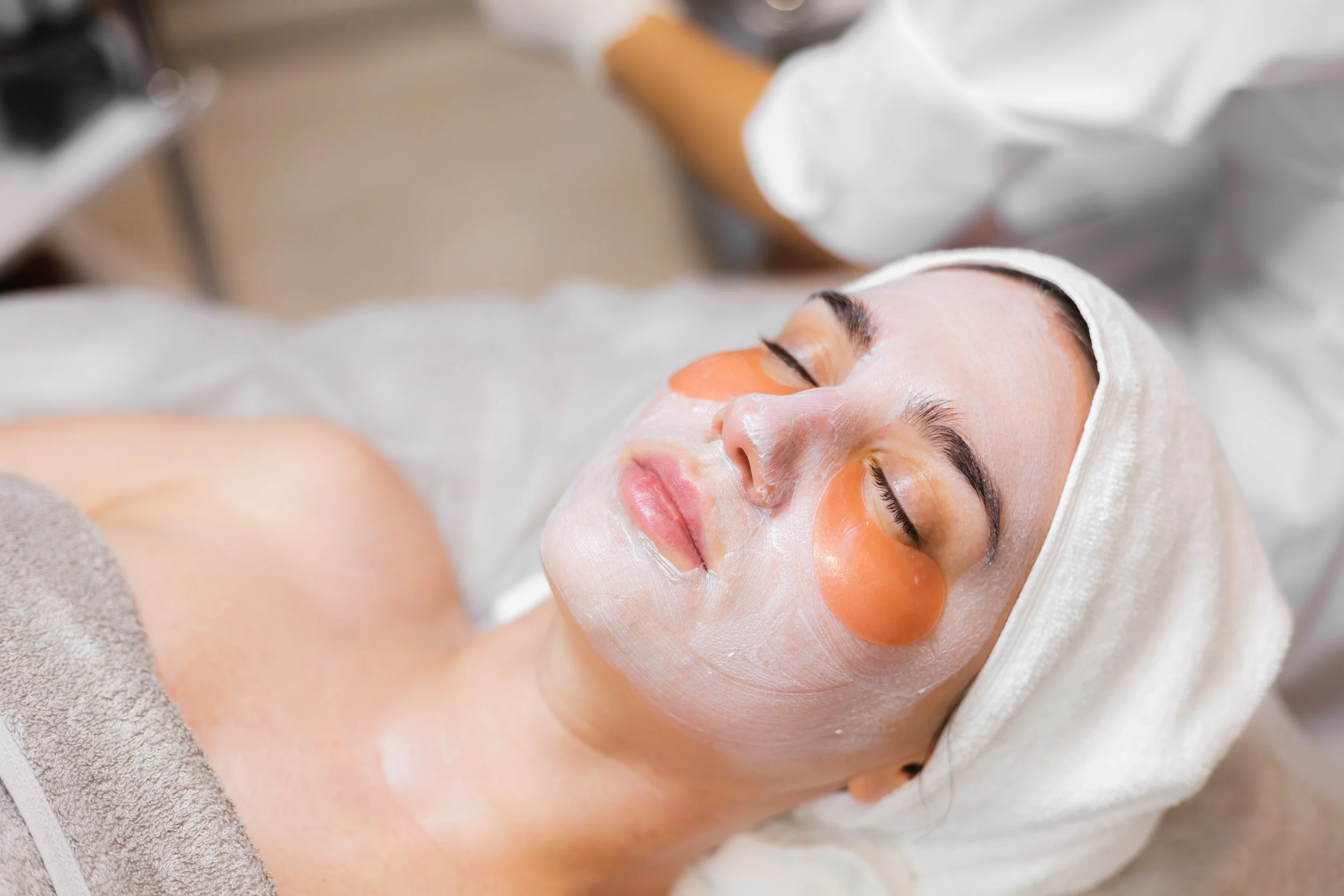 A woman is lying down with her eyes closed, receiving a facial treatment. She has orange eye patches under her eyes, a white facial mask on her face, and a white headband. A skincare professional is applying a treatment or massage to her face in a spa or clinic setting.