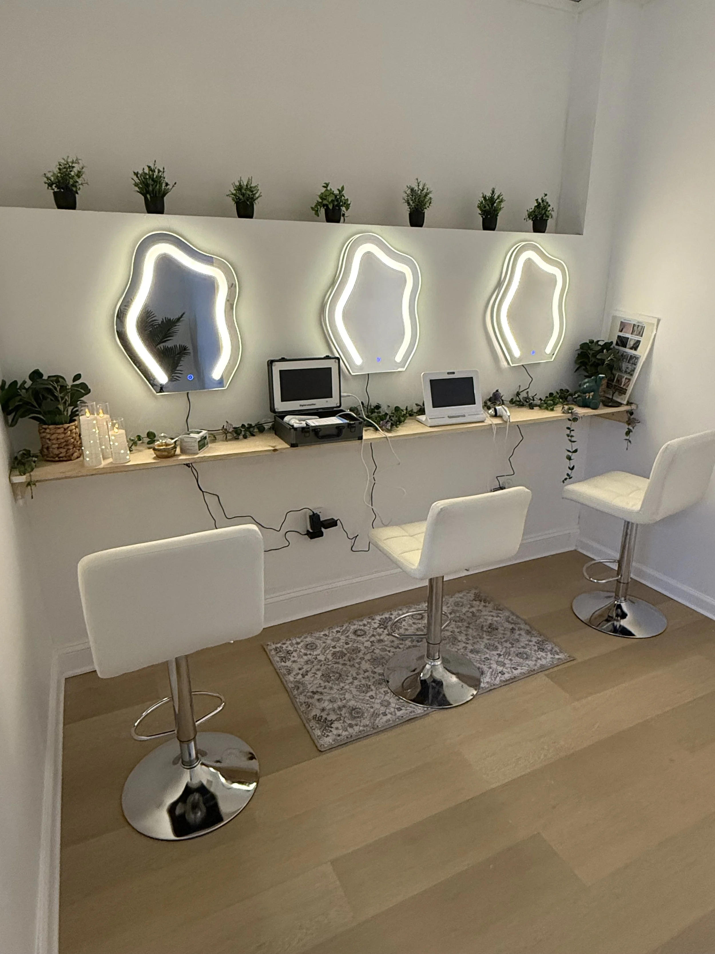 A wall with three illuminated abstract mirror designs, a shelf below with potted plants, electronic devices, candles, and decorative plants, and two white barstools positioned in front.