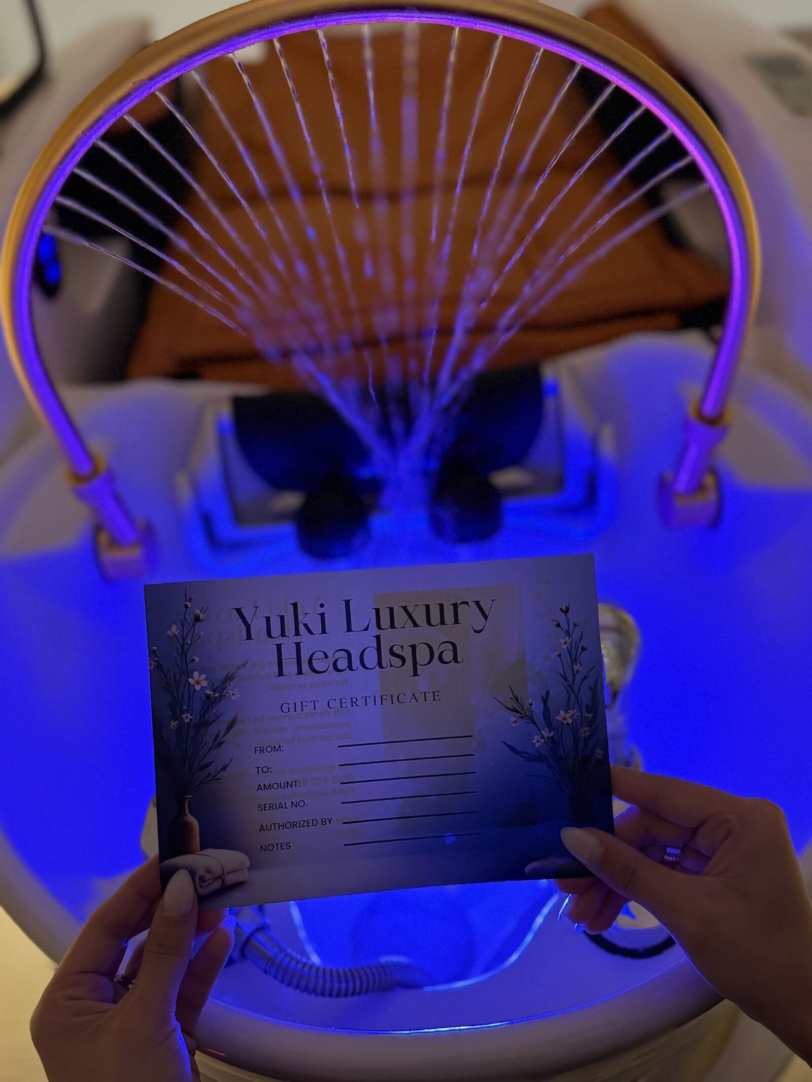 A person holding a gift certificate in front of a glowing machine with purple light, with the certificate reading 'Yuki Luxury Headspa'.
