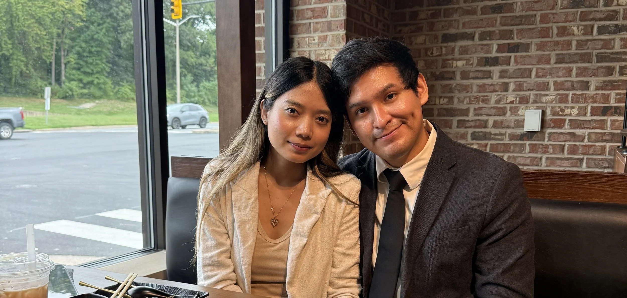 A young Asian woman and a young man are sitting together inside a restaurant, smiling at the camera. The woman has long dark hair and is wearing a beige blazer and a heart-shaped necklace. The man has short dark hair and is wearing a dark suit with a white shirt and black tie. They are sitting in front of a brick wall, near a large window showing some trees and parked cars outside.