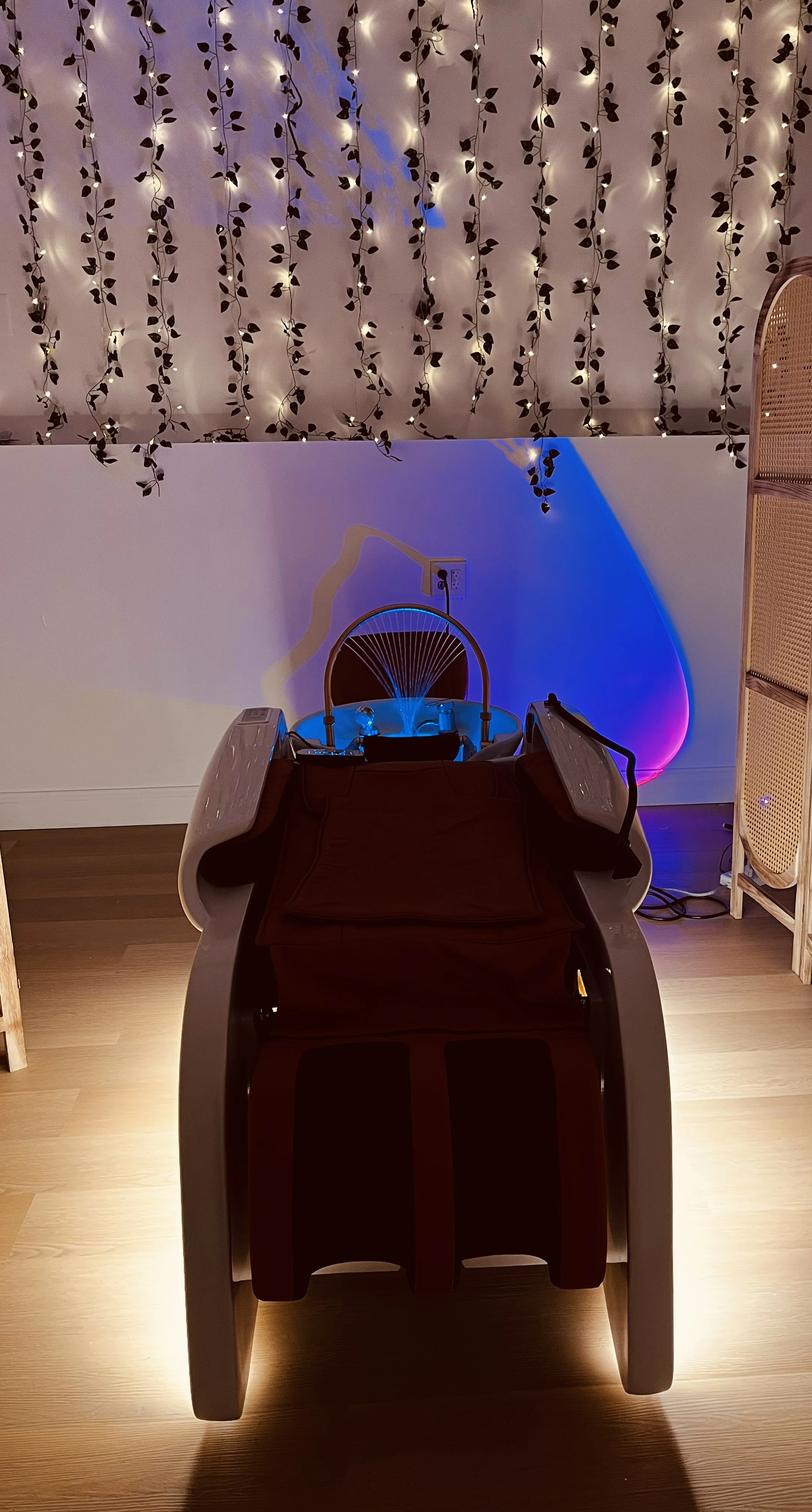 A massage chair with a dark color in a room decorated with hanging string lights and fairy lights on the wall, creating a cozy atmosphere.