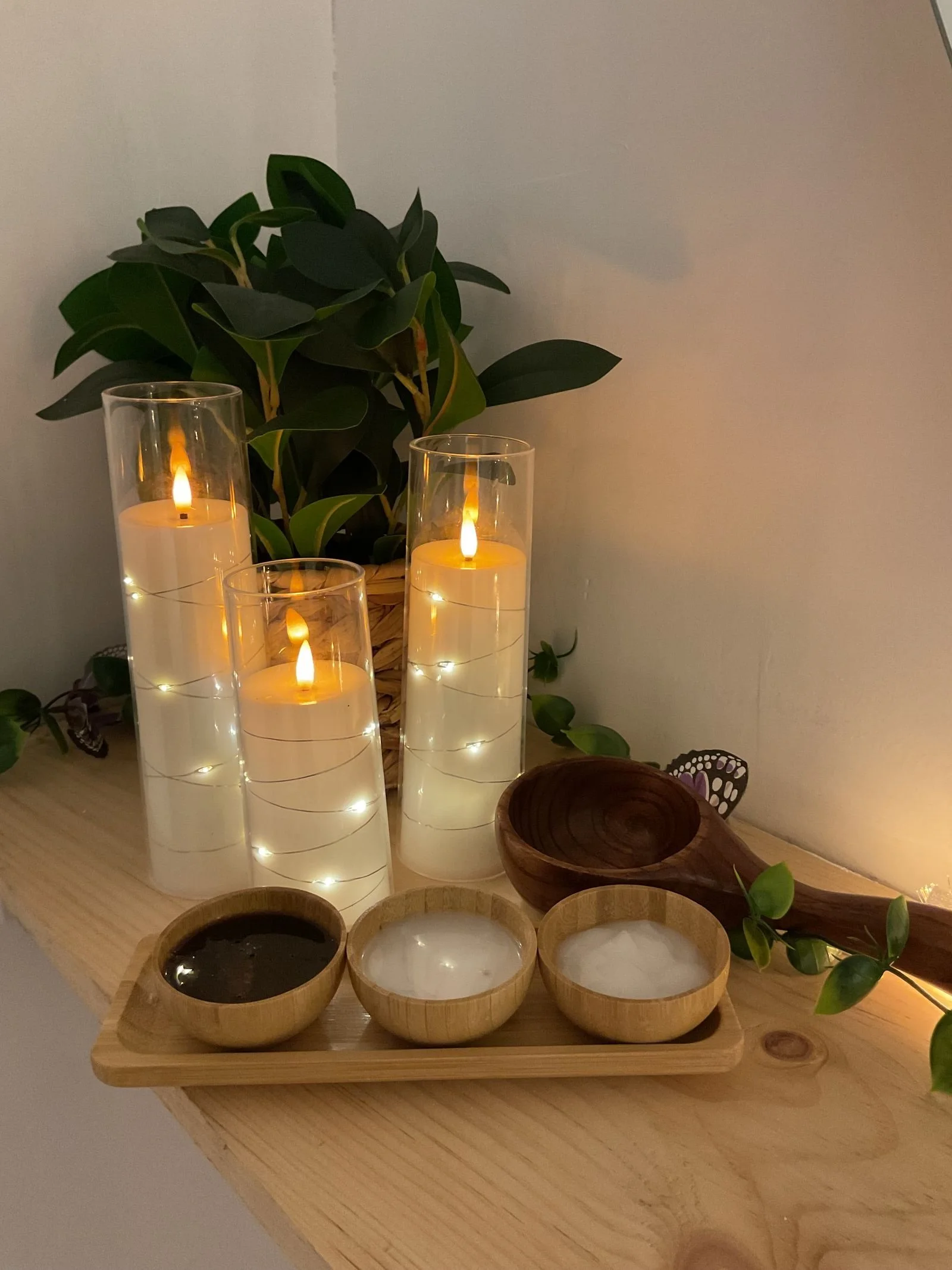 Decorative setup with three glass candles with flickering flames, a green leafy plant, and wooden bowls on a wooden surface.