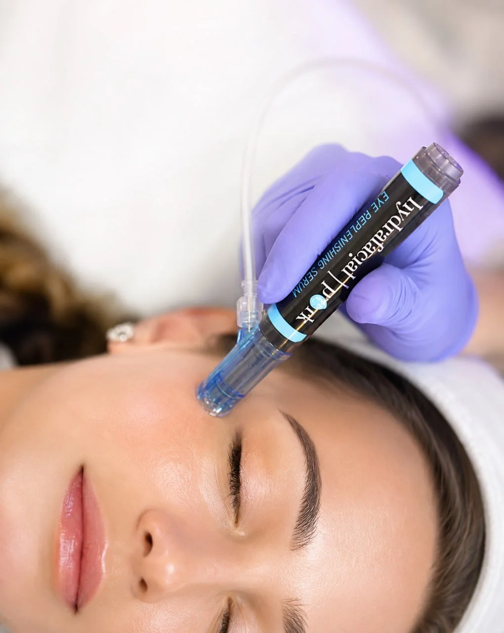 A woman receiving a facial treatment with a serum injection in a clinical setting, lying with her eyes closed while a practitioner administers the procedure.