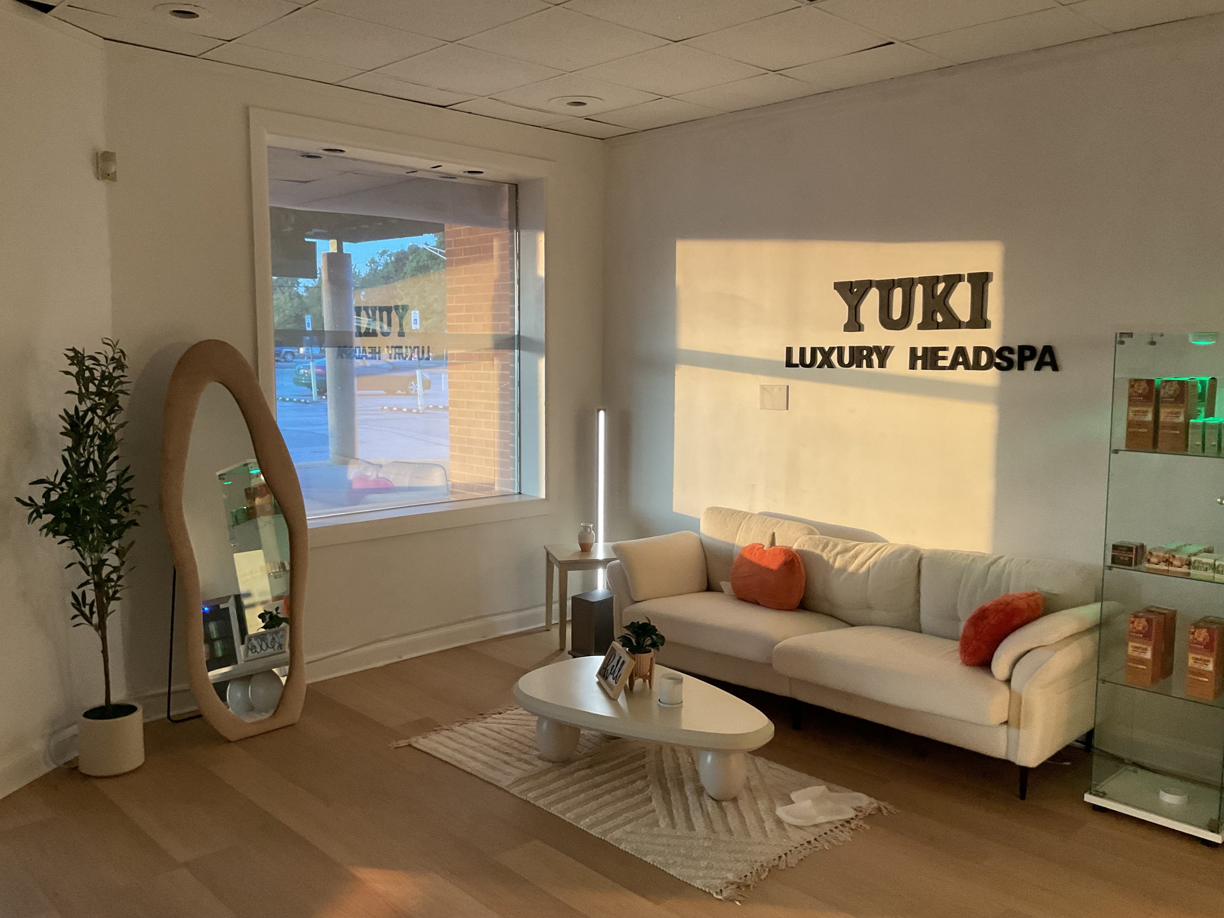 Interior of a lounge area at a luxury headspa with a white couch decorated with orange pillows, a round white coffee table, a tall potted plant, and a large window showing the outside view.