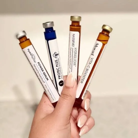 Hand holding five small vials with labels for skincare serums or medical ampoules, each with a different colored cap.