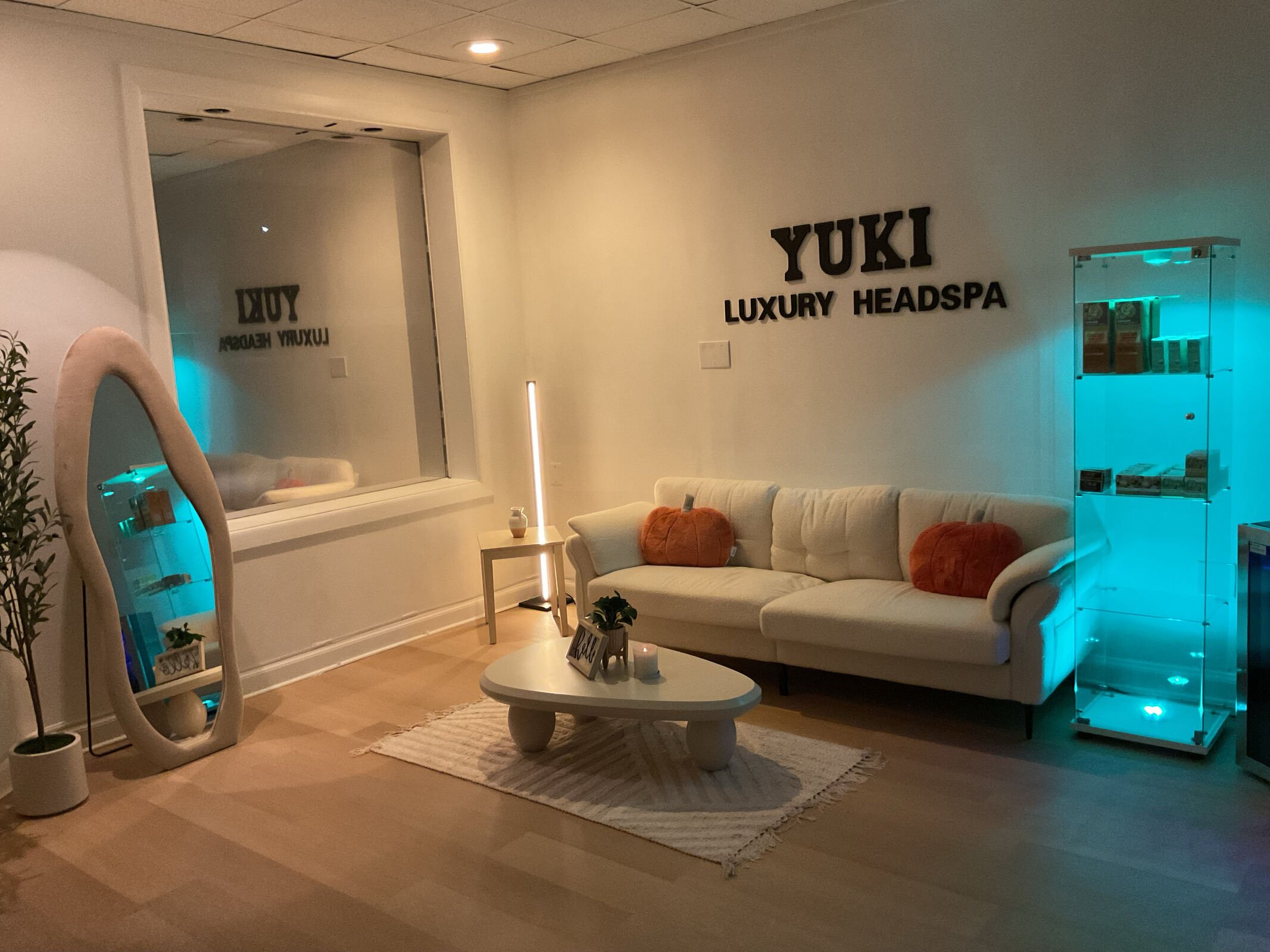 A cozy lounge area with a cream sofa, orange cushions, a small white coffee table, and a patterned rug. There is a white side table with a decorative item and a small potted plant. The wall displays the words 'YUKI LUXURY HEADSPA' in large black lett