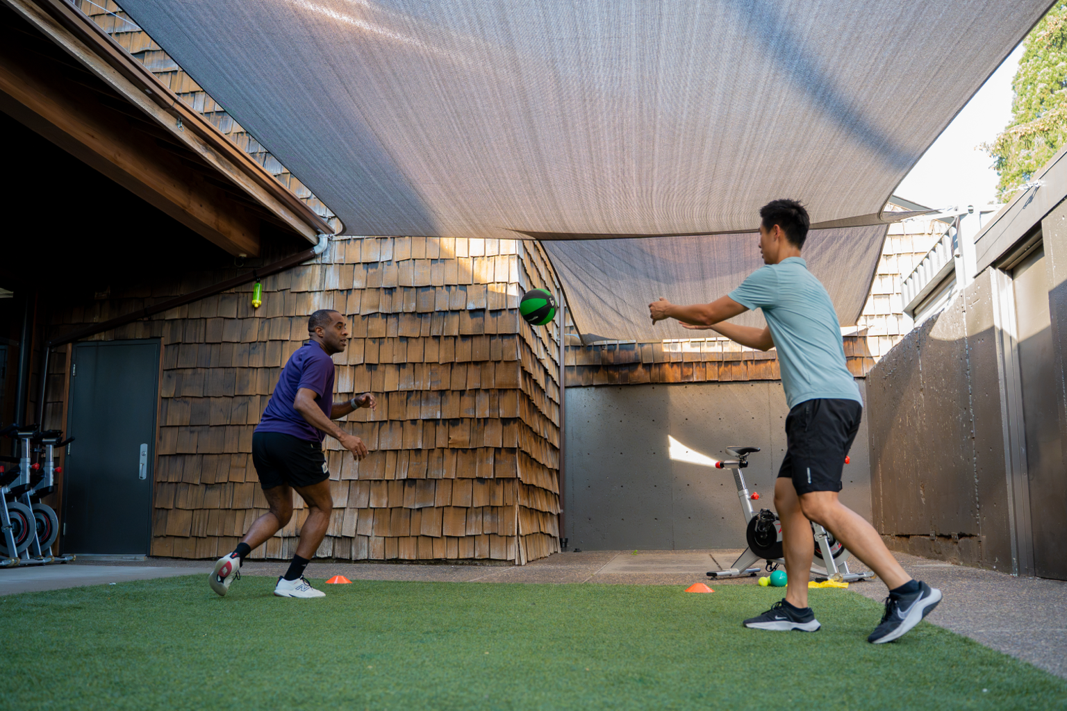Two men playing with a green and black volleyball in a backyard with artificial grass and a purple athletic shirt, a blue shirt, black shorts, a bicycle, and cones for a game or exercise.