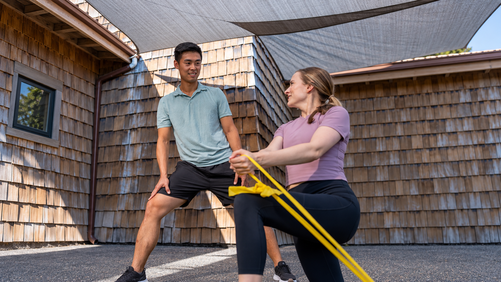 A woman is doing a resistance band exercise outdoors while a man stands nearby. They are on a paved surface with wooden building siding and a shade cloth overhead.