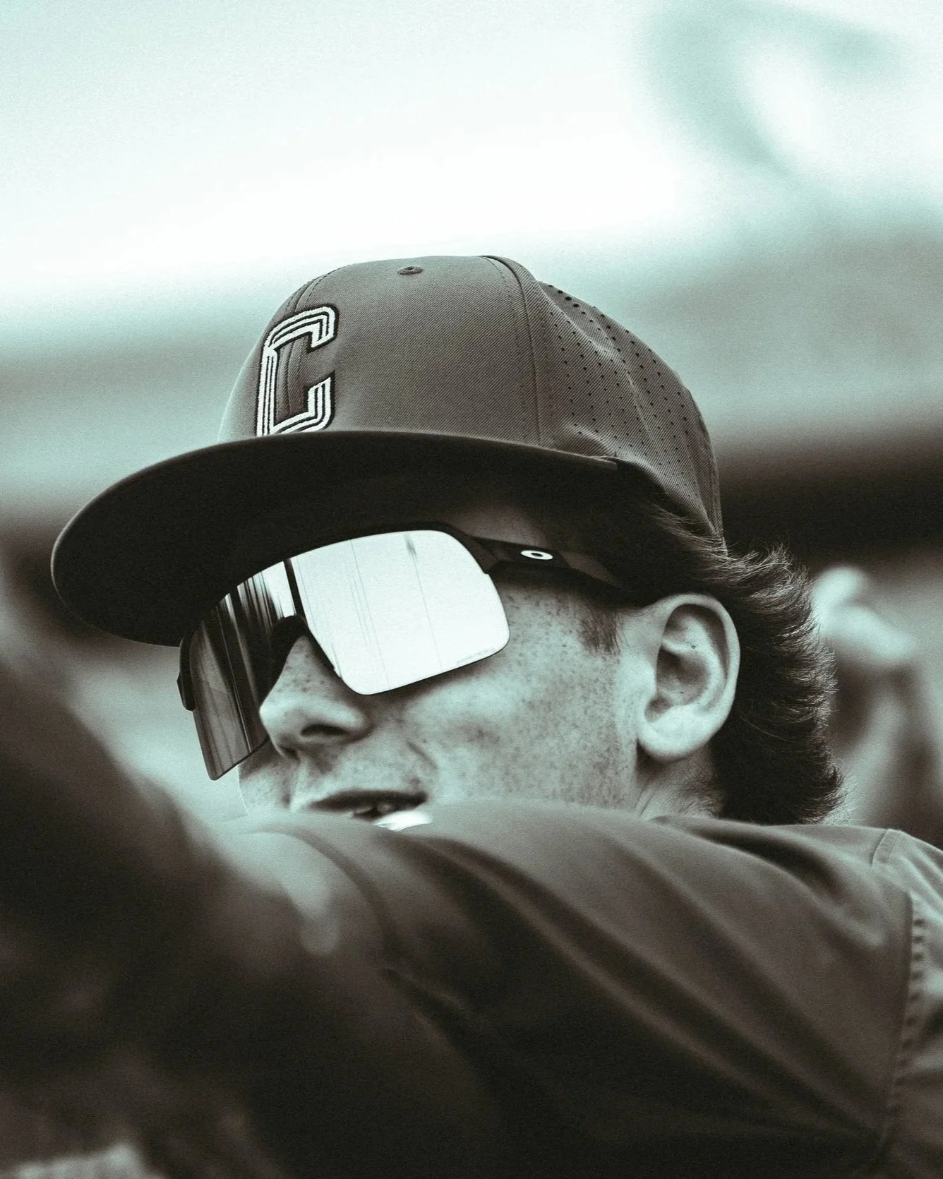 A person with wavy hair wearing a cap, sunglasses, and a jacket, captured from below inside a vehicle with a blurred dashboard.