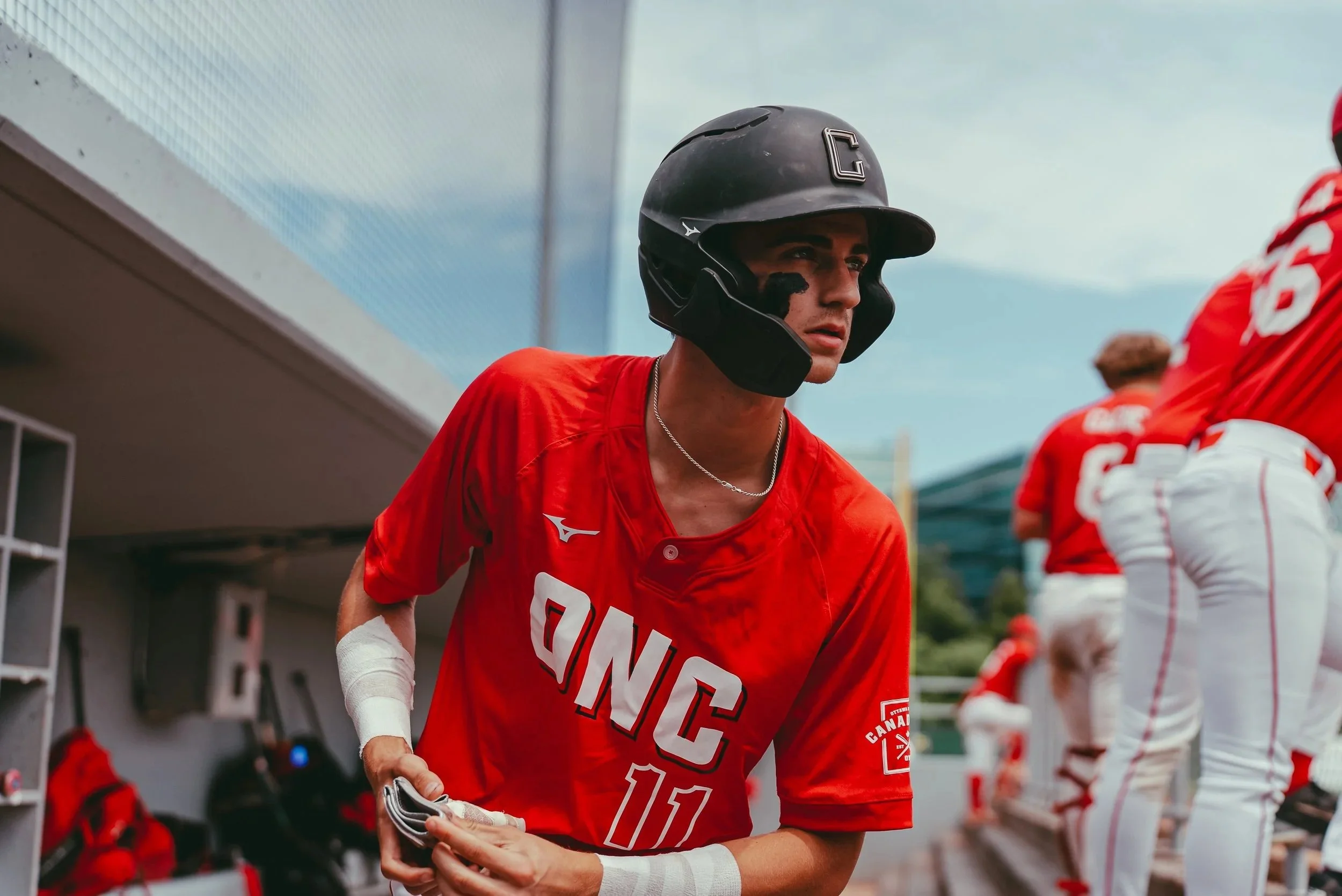 A young male athlete in a red sports jersey and black helmet stands near a dugout during a game, with other team members in the background.