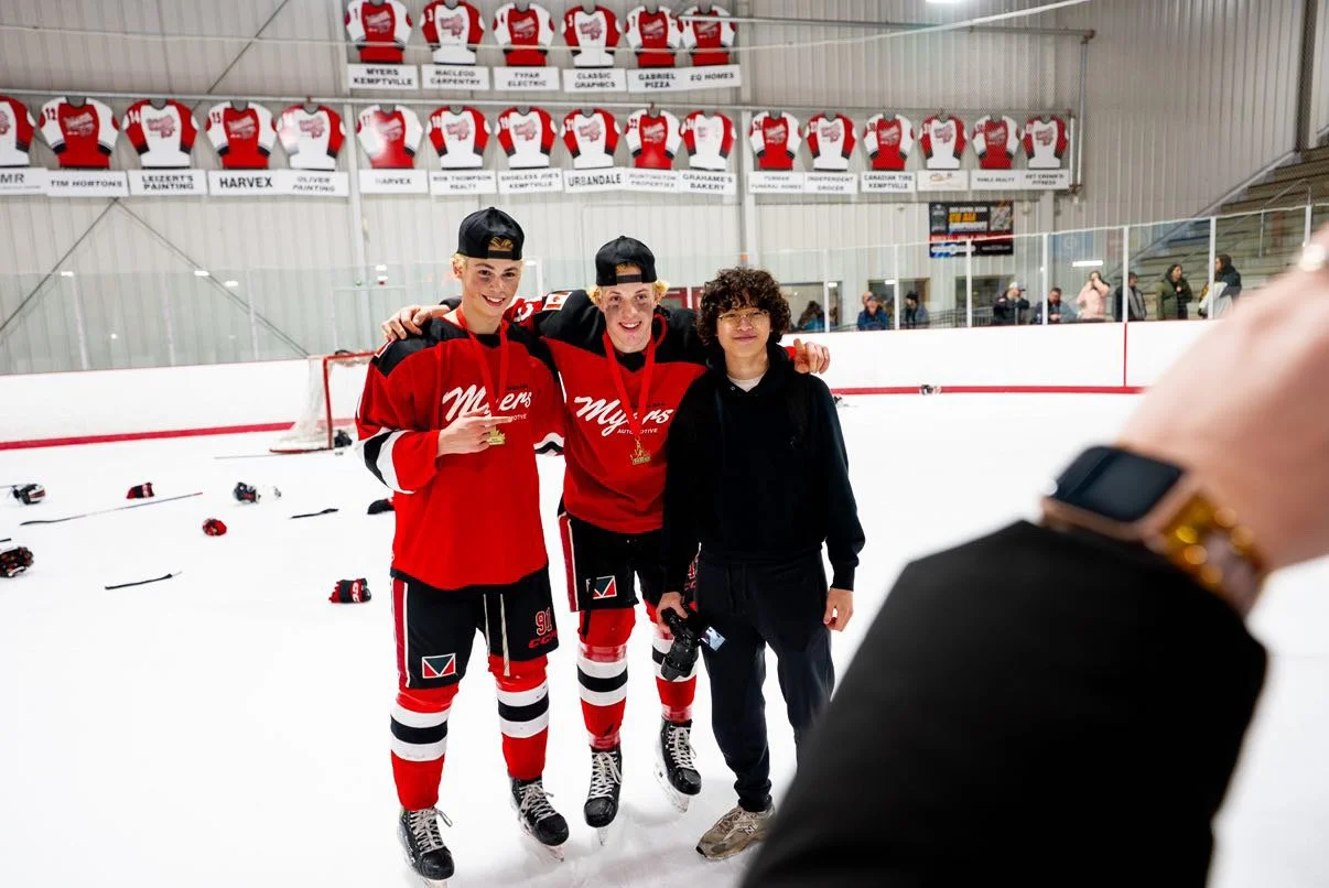 Three people standing on an ice hockey rink with two wearing red hockey jerseys and medals, and one in casual clothes, with hockey gear and jerseys hanging on the wall behind them.