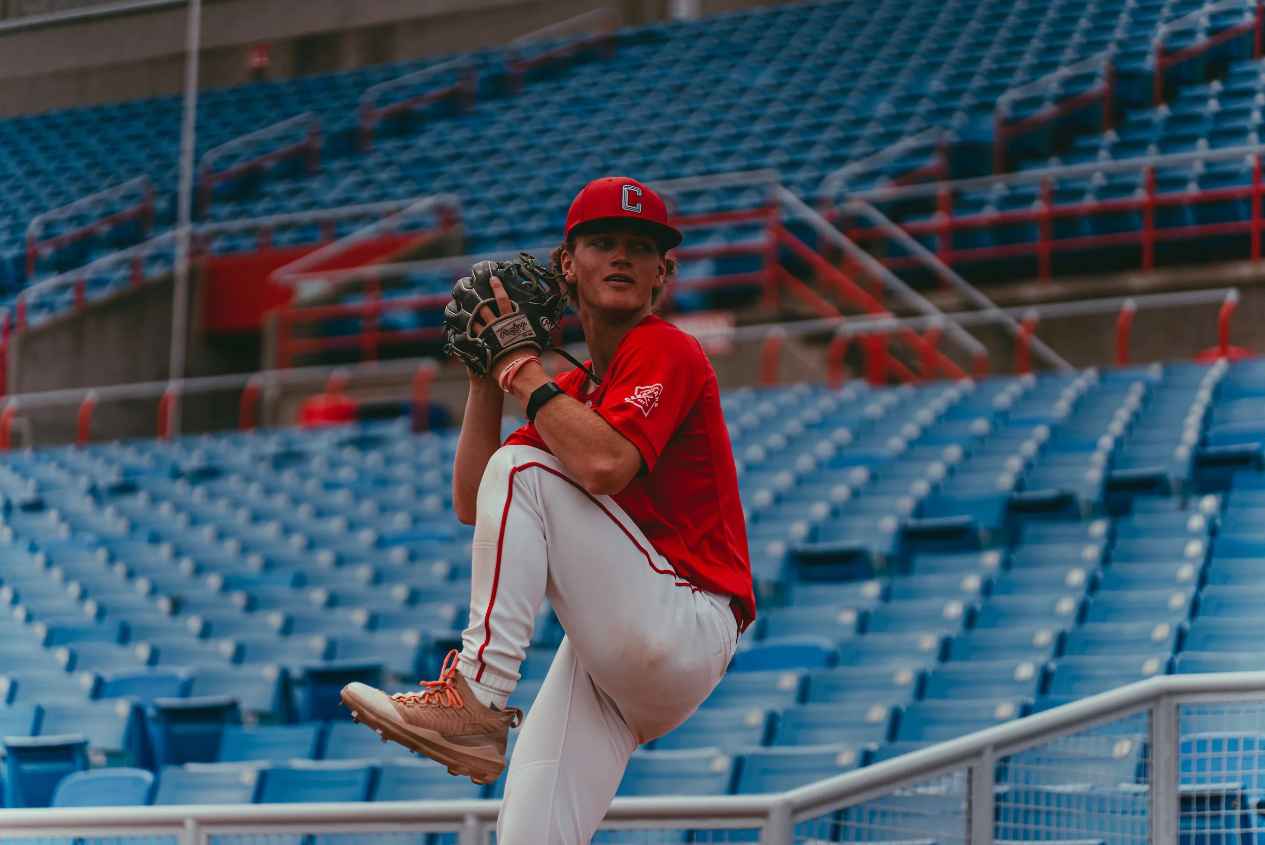 A young male baseball player in red and white uniform doing warm-up stretches in empty stadium seats.