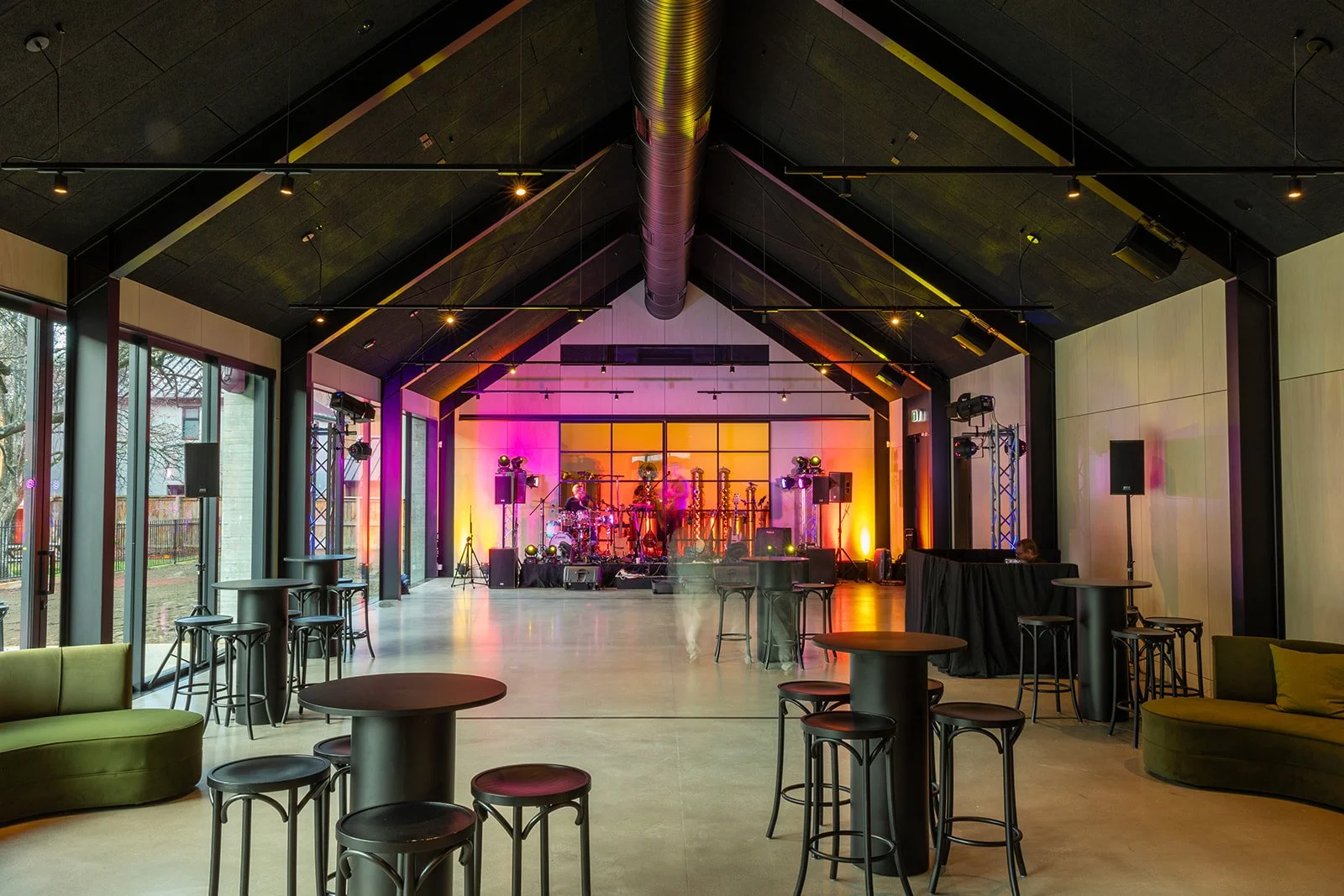 Empty indoor venue with a stage set for a live music performance, colorful lighting, and various seating arrangements including high tables, bar stools, and green couches.