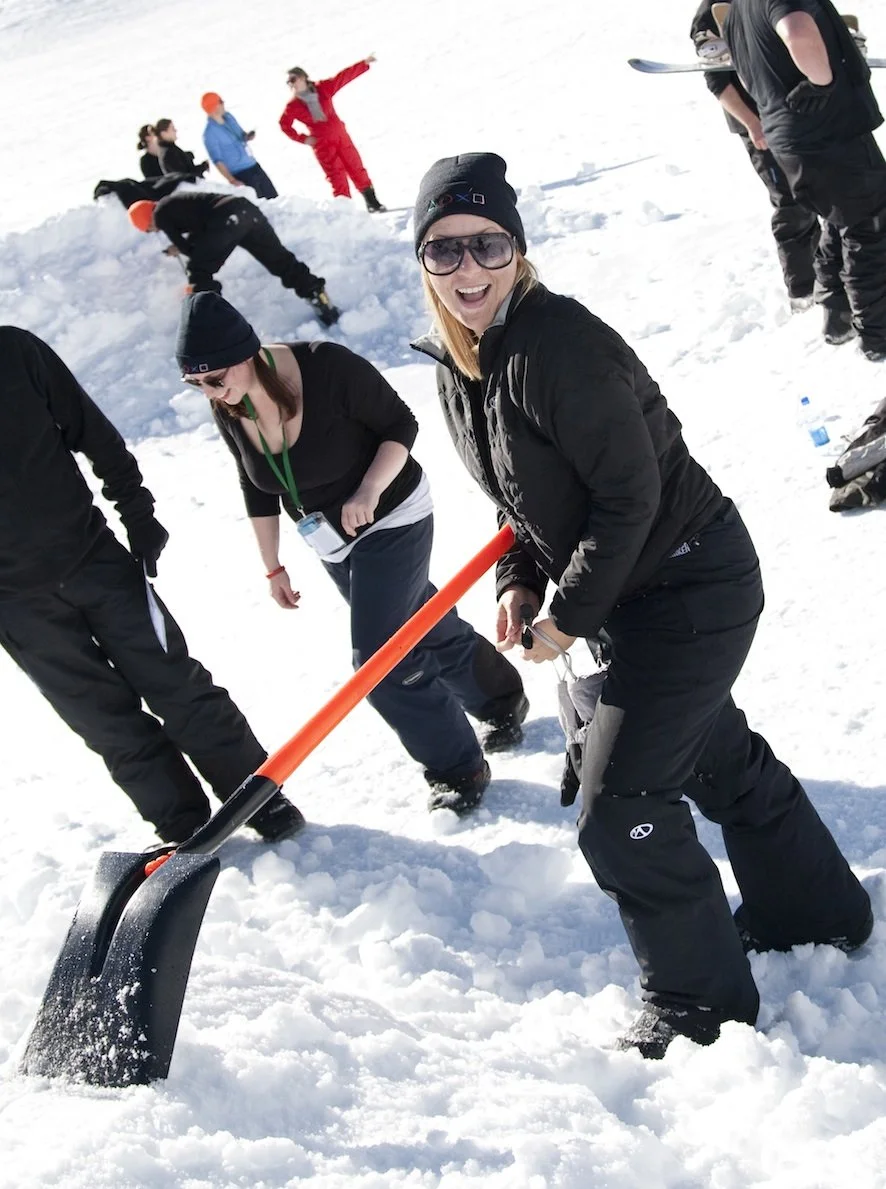 People enjoying a snowy day, with one woman smiling and holding a snow shovel, others in the background playing and walking on snow. Outdoor team building in the snow.