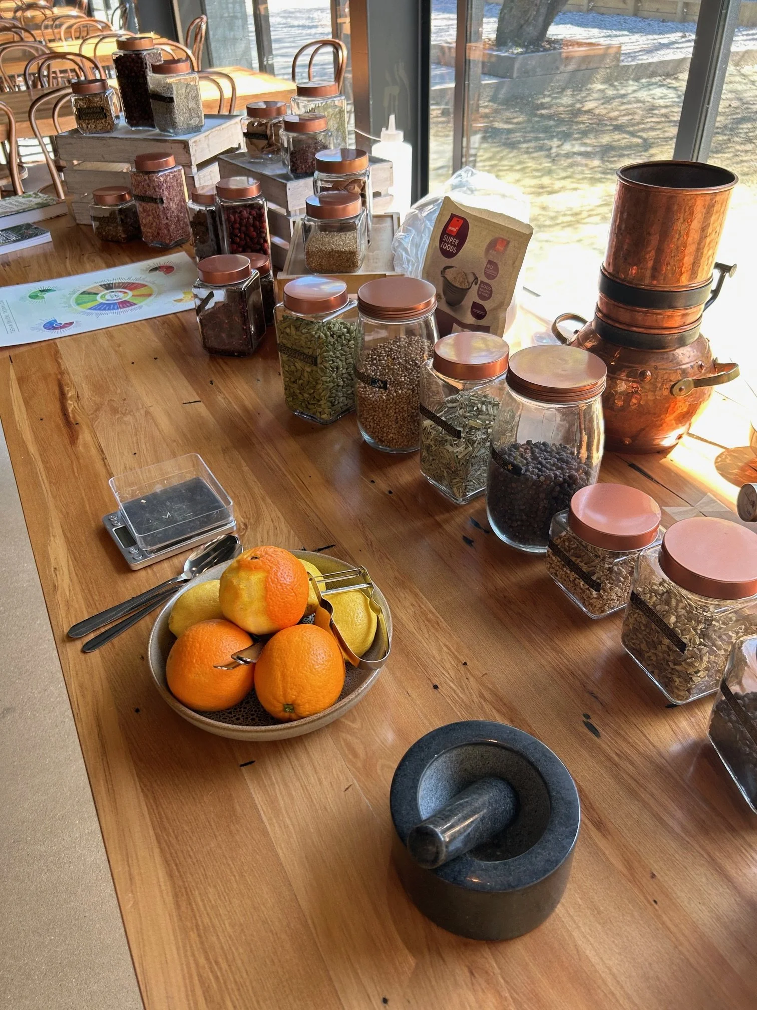 Indoor team building. Create your own gin workshop. A wooden table with glass jars of various spices and herbs, a bowl of oranges and lemons, a meat grinder or mortar and pestle, and a coffee filter machine near a window with sunlight streaming in.