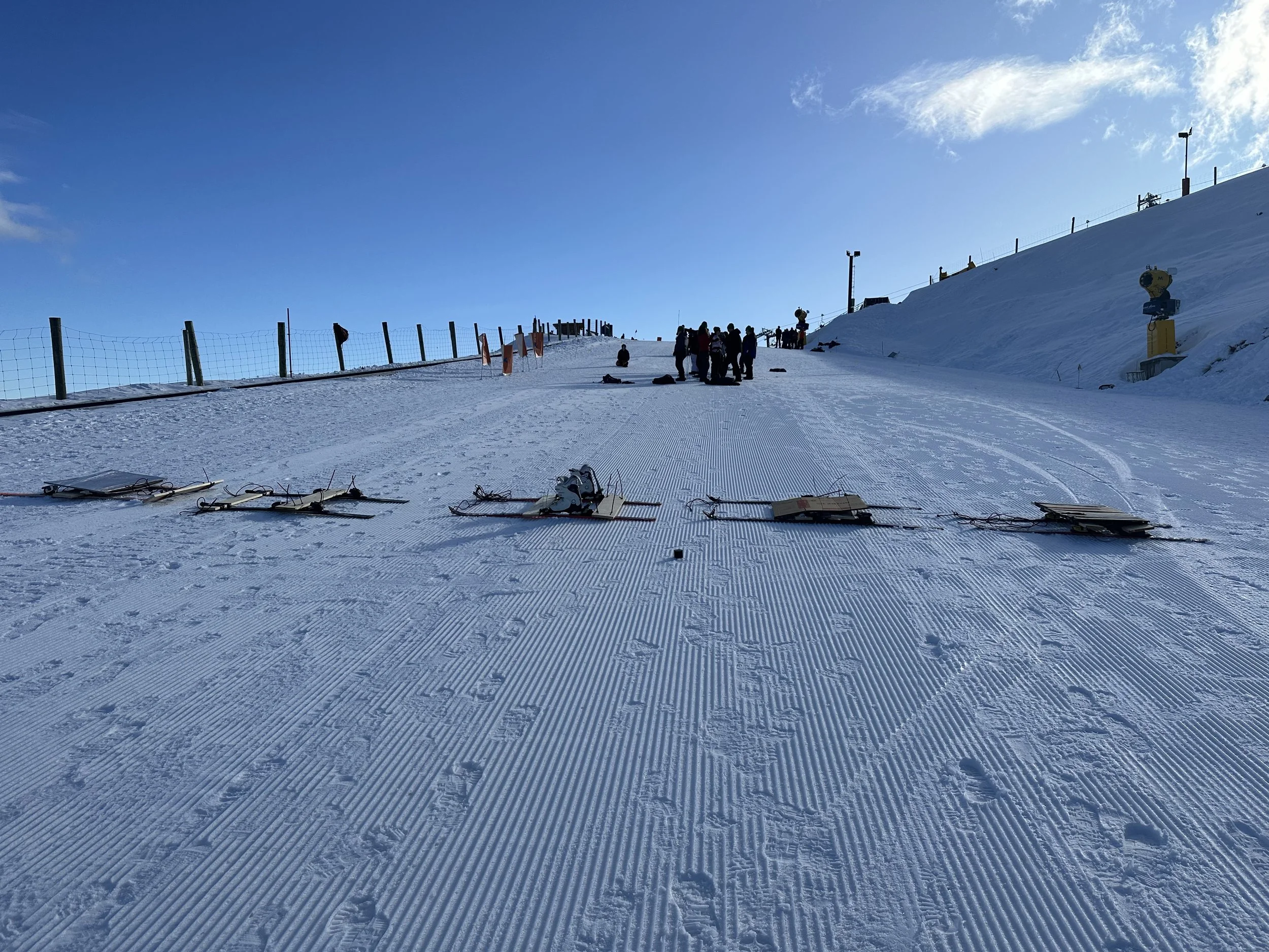 Snow-covered ski slope with ski equipment on the ground, group of people in the distance, and ski lift structures against a clear blue sky.