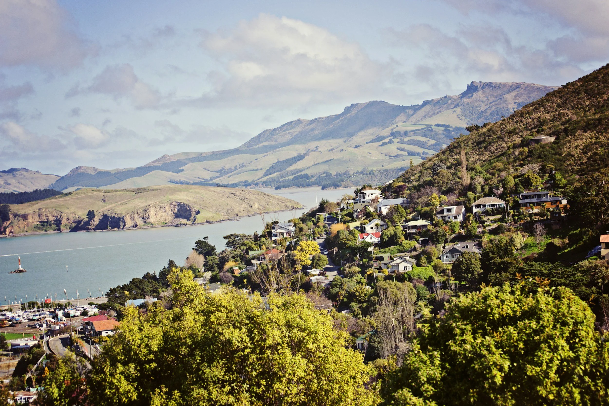 A scenic view of a small town with houses and trees on a hillside beside a body of water, with mountains in the background under a partly cloudy sky.