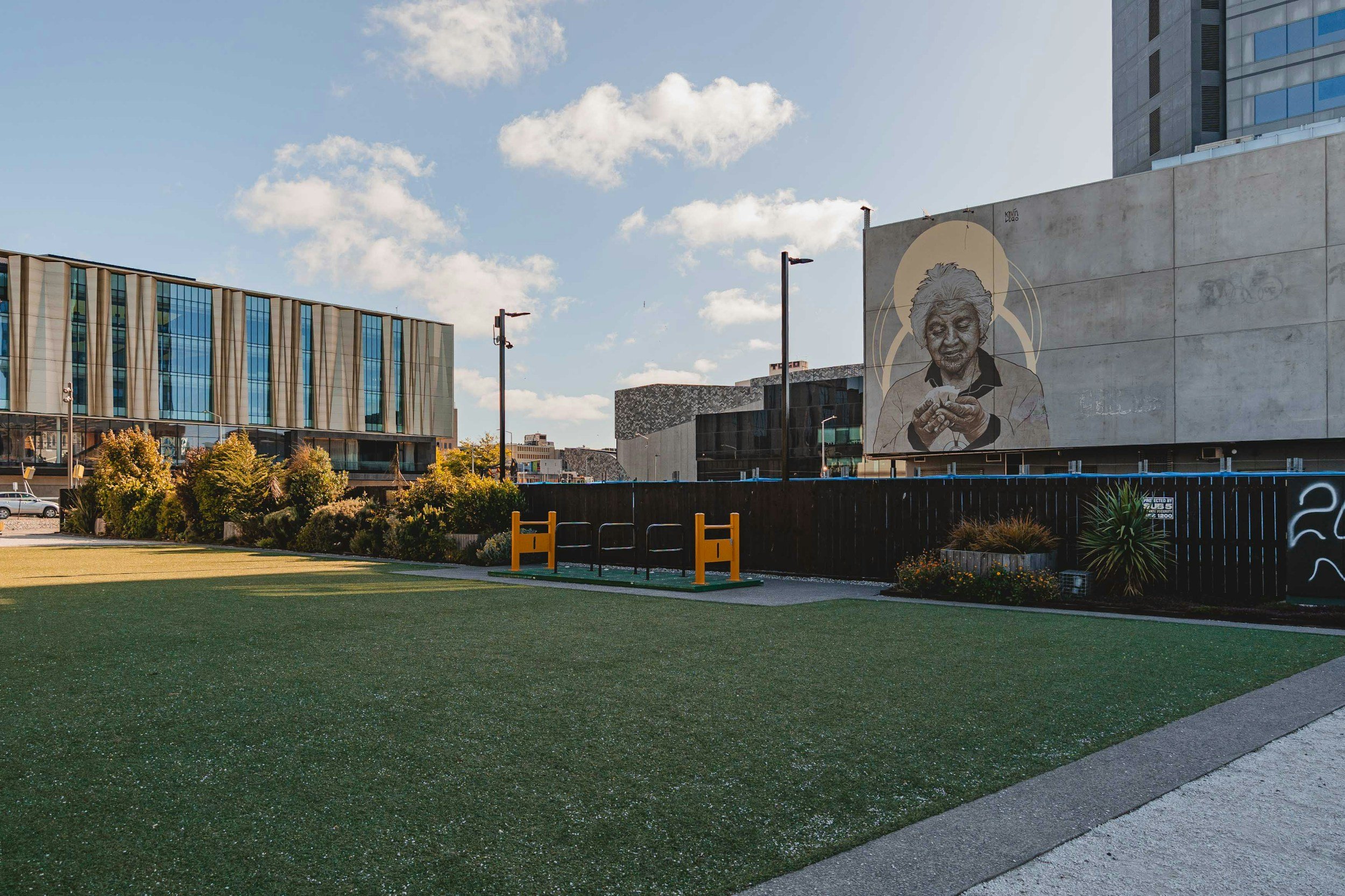 Cityscape with modern buildings, street lamp posts, trees, a grassy area, and a mural of an elderly woman on a concrete wall.