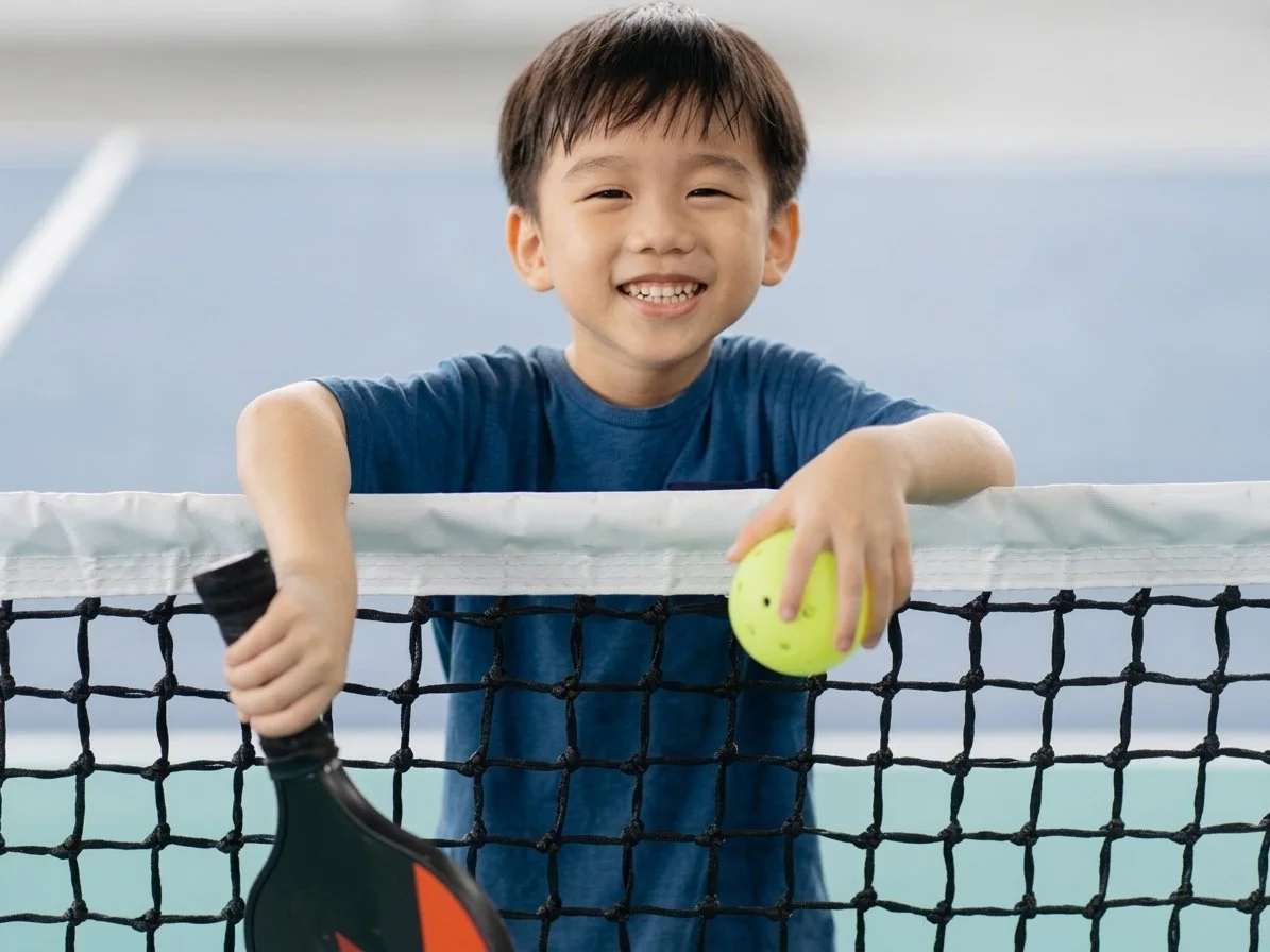A smiling boy with short brown hair wearing a blue t-shirt holding a tennis racket and tennis ball behind a tennis net.