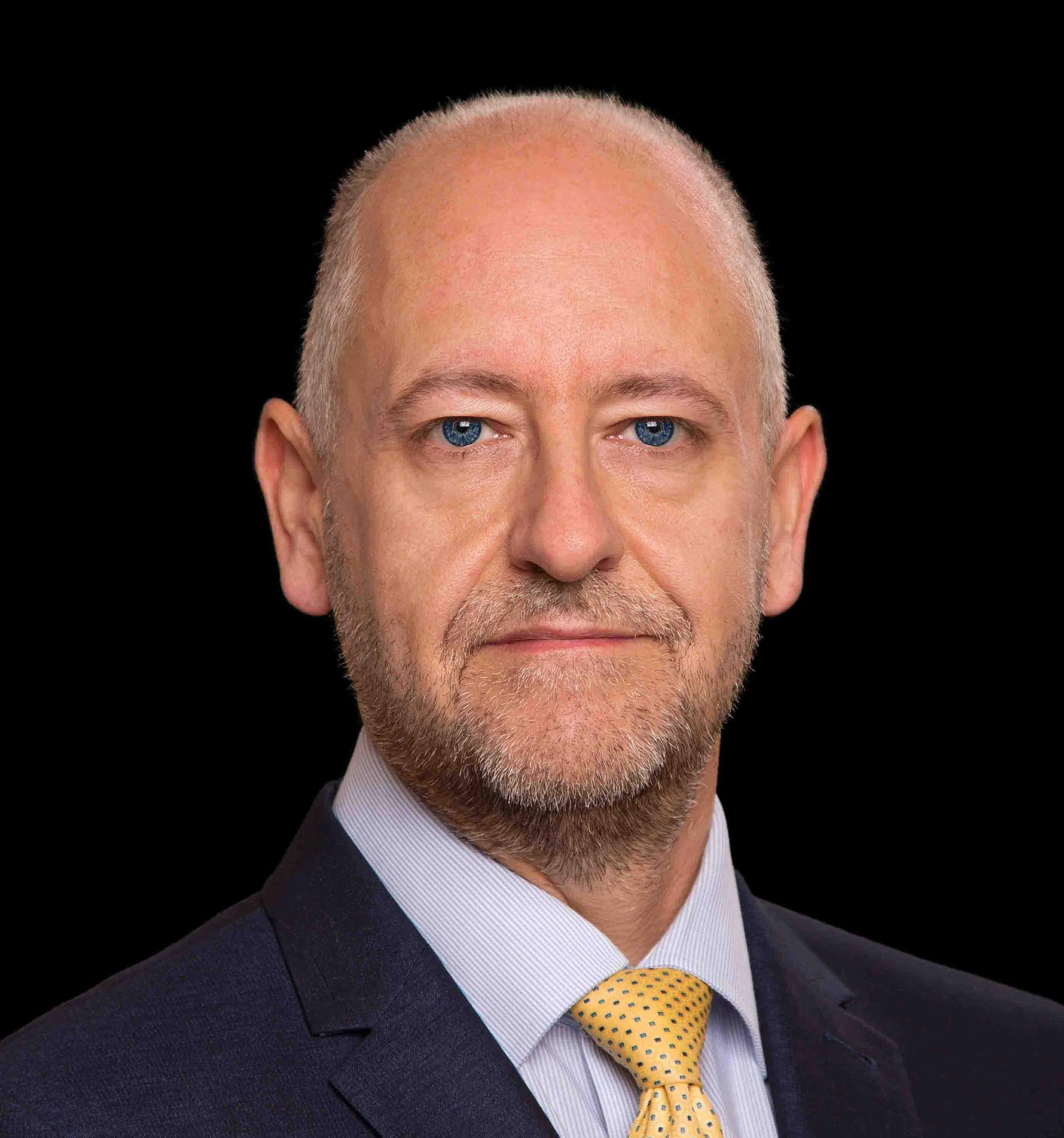 Close-up of a middle-aged man with blue eyes, grey hair, and glasses, dressed in a navy suit, white shirt, and yellow tie with blue dots, against a black background.