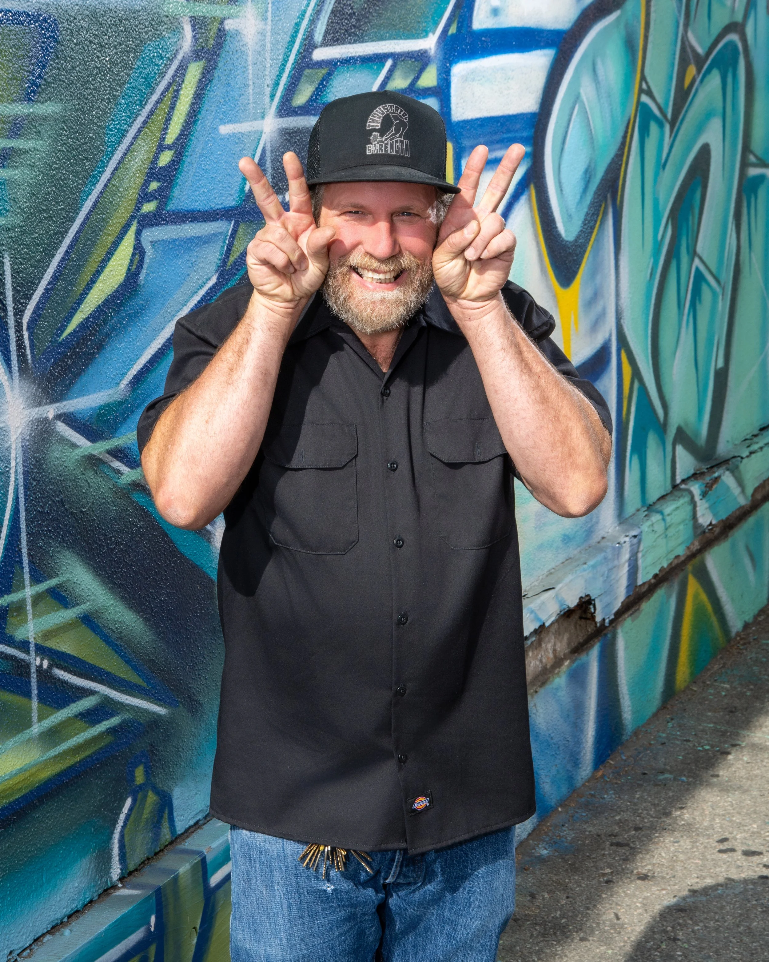 A smiling man with a beard wearing a black cap and black shirt, making a peace sign with both hands, standing in front of a colorful graffiti wall.