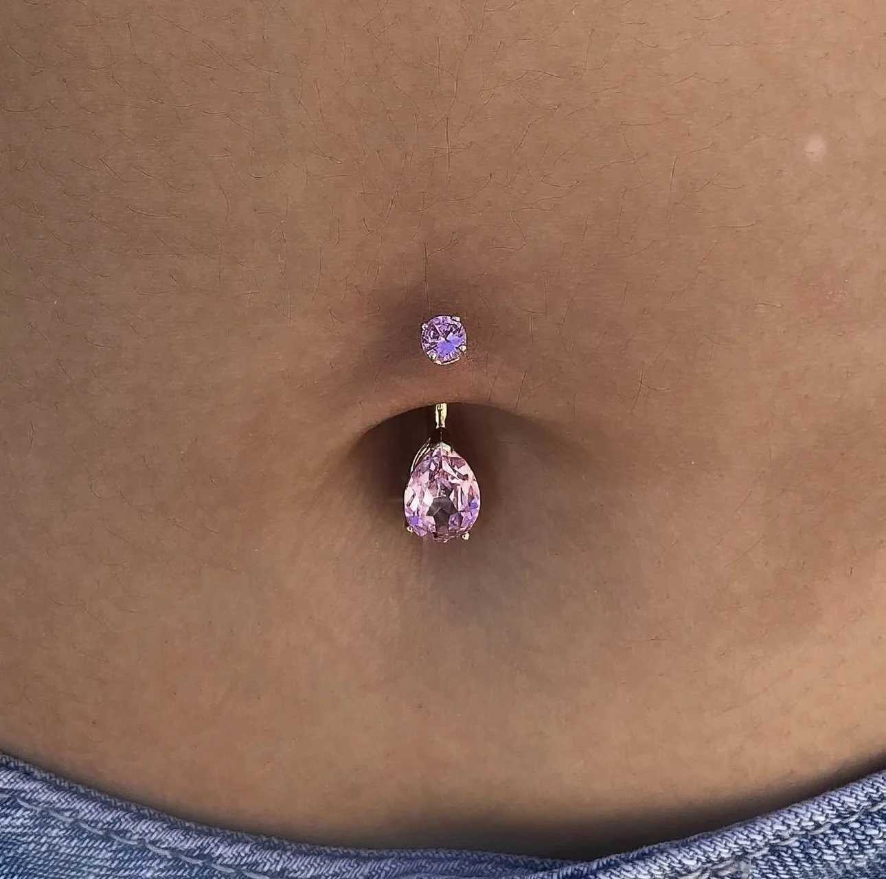Close-up of a person's navel with a two-piece pink and purple gemstone belly piercing.