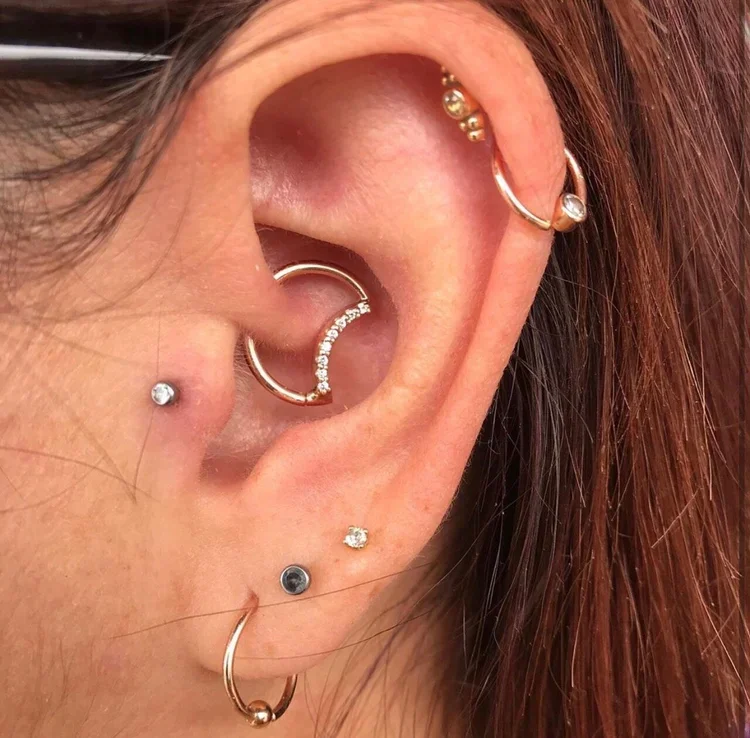 Close-up of a person's ear adorned with multiple piercings and jewelry, including hoop earrings, stud earrings, and a curved barbell, with red hair visible.