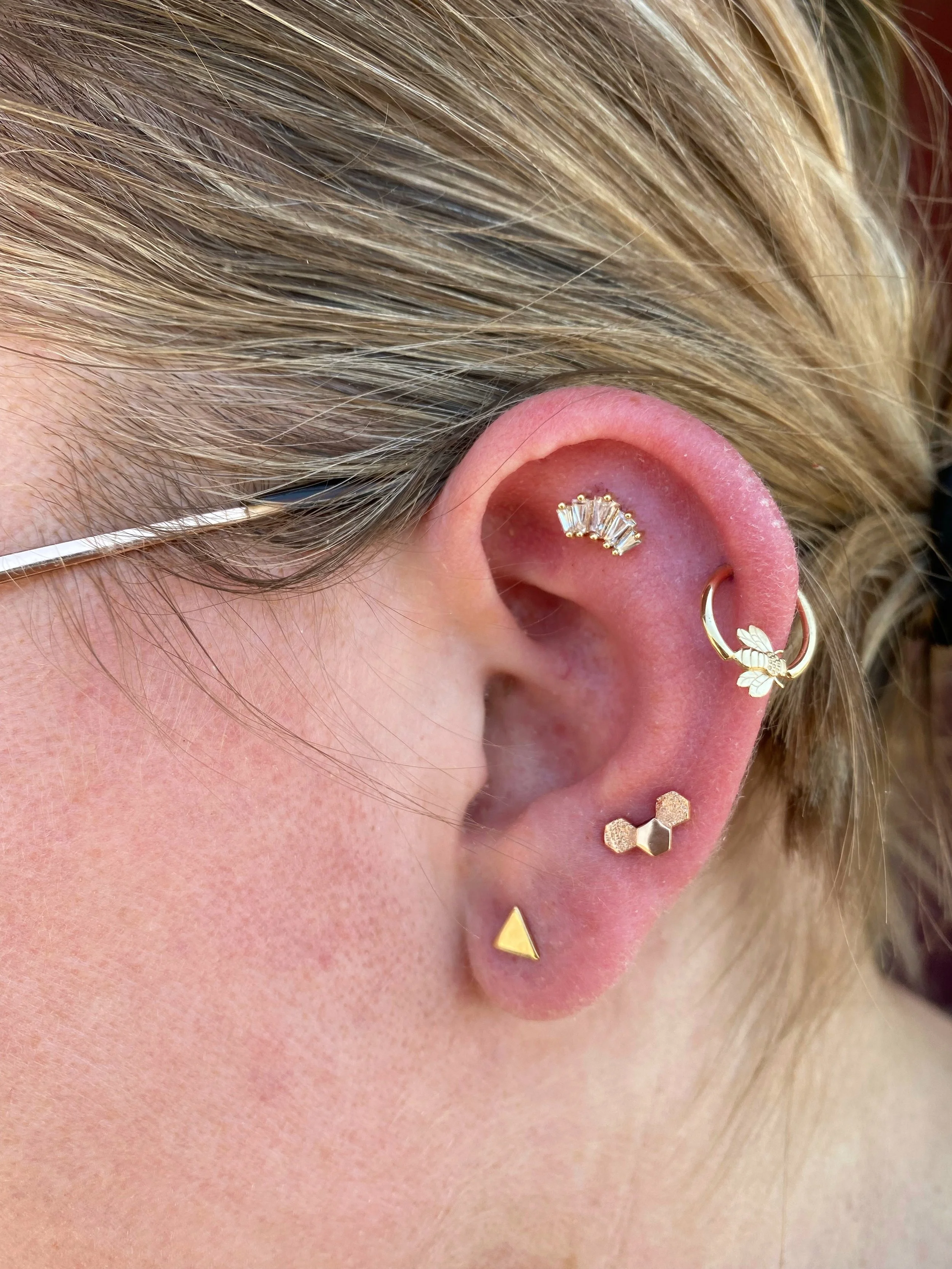 Close-up of a person's ear with multiple gold earrings, including a curved cartilage earring with clear gemstones, a small hoop with a leaf charm, and other geometric studs, and short blonde hair.