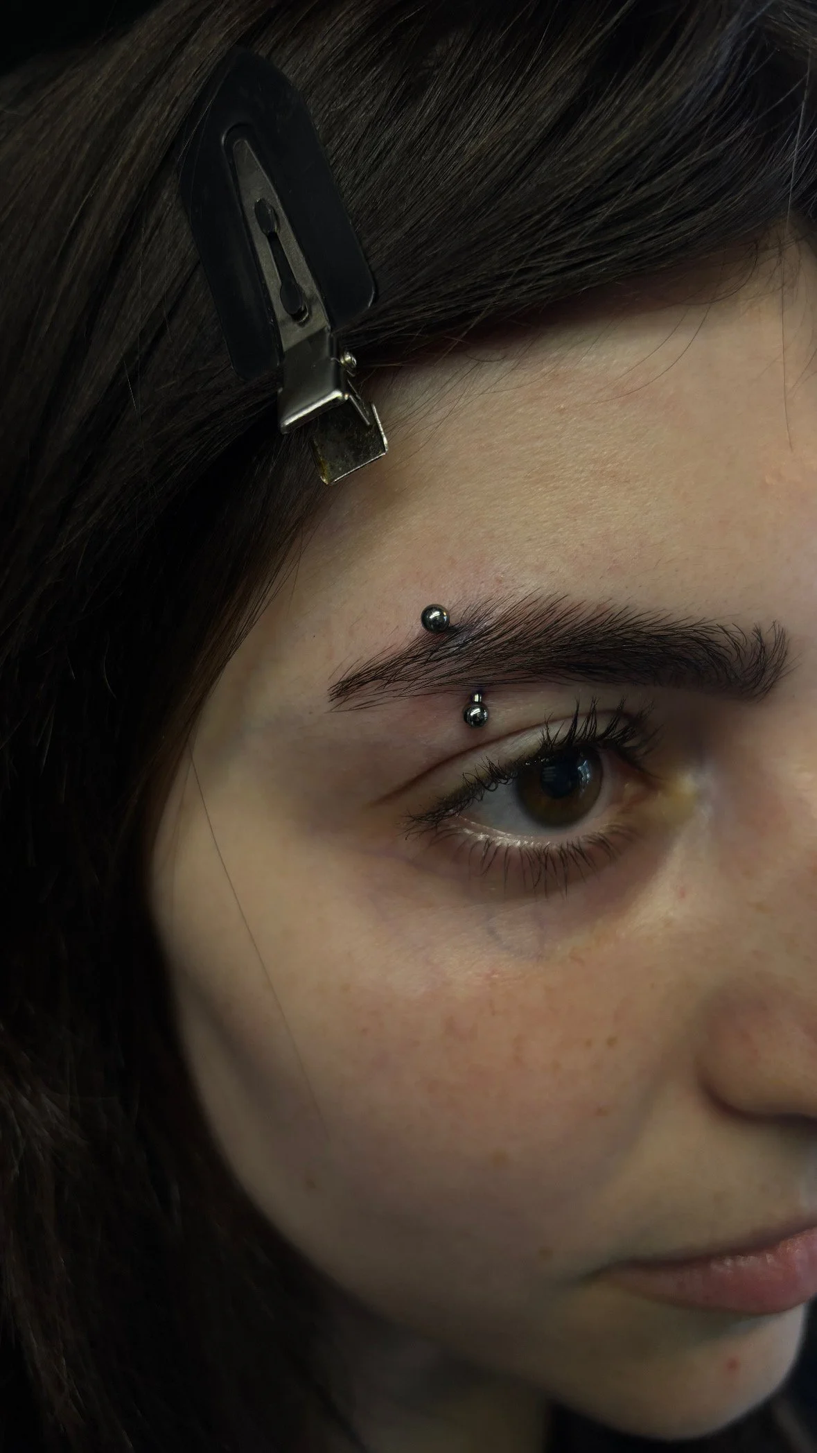 Close-up of a young woman's face with a facial piercing above her right eyebrow, dark hair held back with a hair clip, and a slight smile.