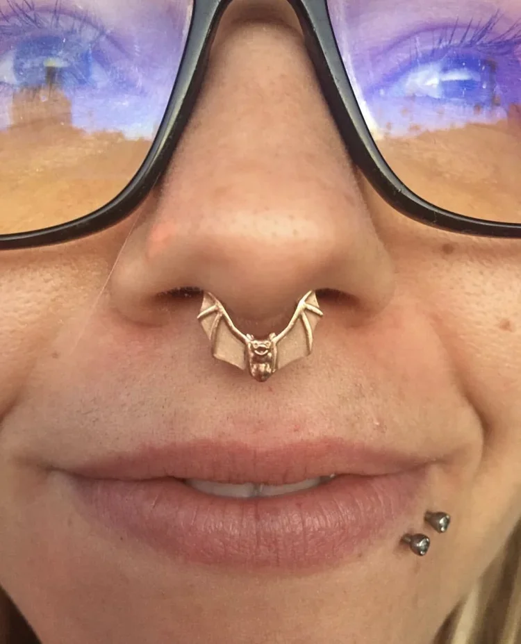 Close-up of a person's face with glasses, a bat-shaped nose ring, and two lower lip piercings, smiling slightly.