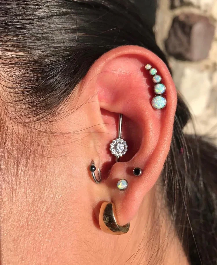 Close-up of a person's ear with multiple earrings, including a safety pin with a dangling decorative element, a crystal hoop, a small black stud, a pearl-like stud, and a gold hoop.