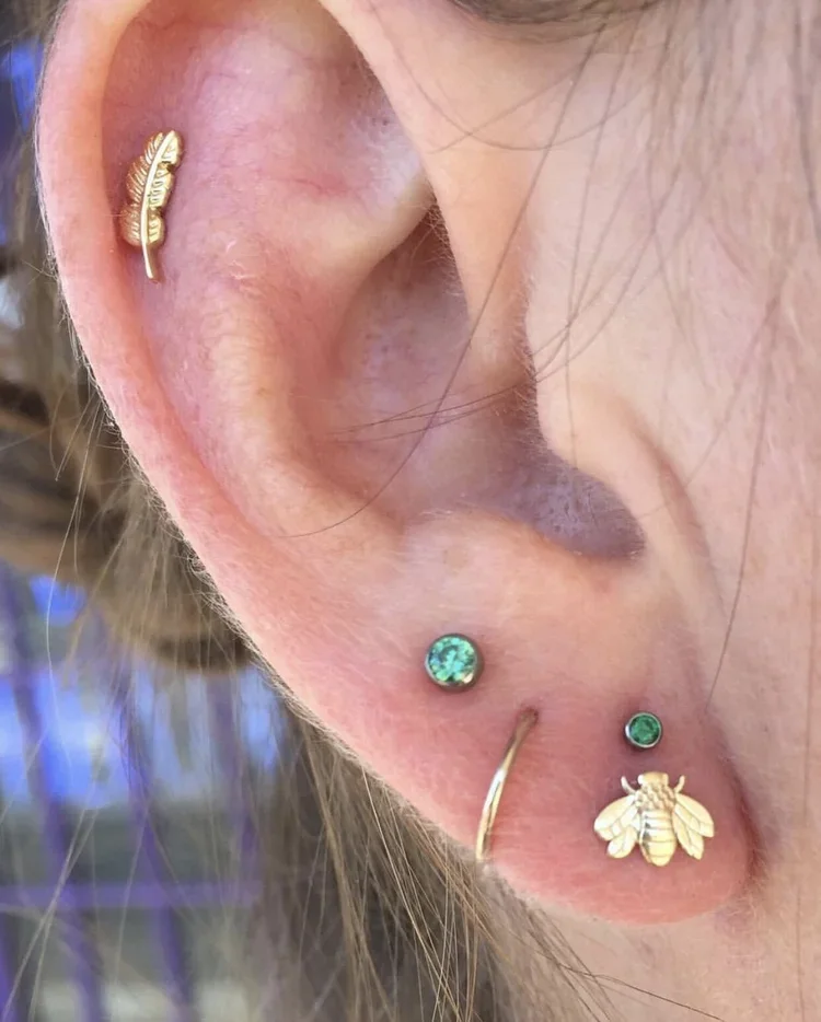Close-up of a person's ear with multiple piercings, decorated with various earrings including a bee, green gemstones, and a gold hoop.