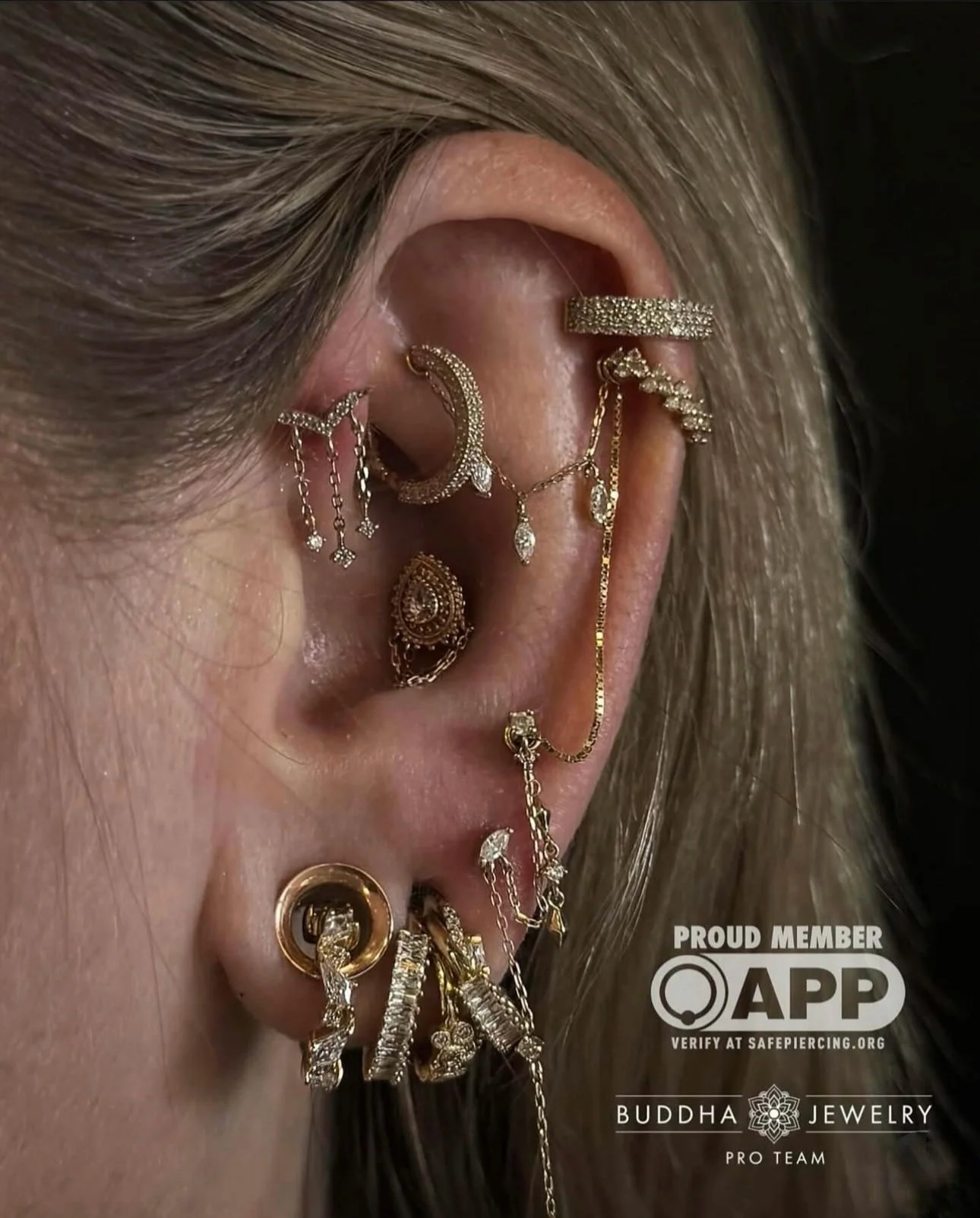 Close-up of a person's ear filled with gold and silver earrings, chains, and decorative jewelry, including rings and ear cuffs from the Buddha Jewelry Collection