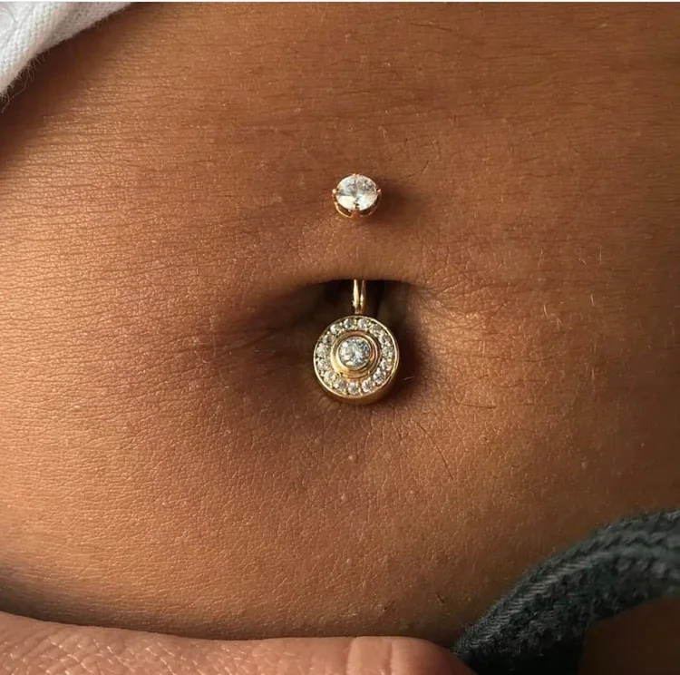 Close-up of a belly button piercing with a round gemstone on top and a circular pendant with a gemstone in the center surrounded by smaller gemstones hanging below.