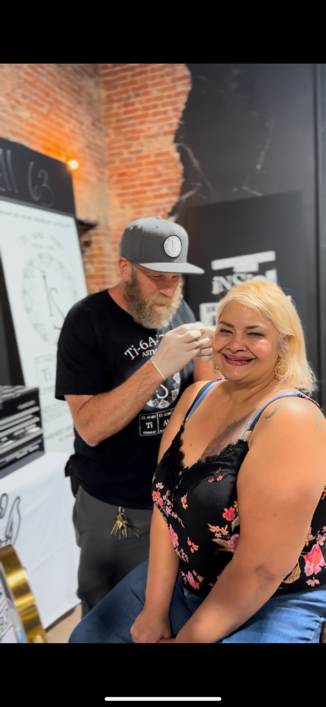 A woman receives a tattoo on her arm by a tattoo artist in a room with brick walls. The woman is smiling, and the artist is focused on his work.