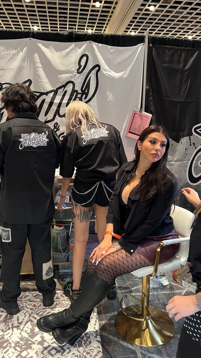 A woman with long dark hair seated on a gold and white chair at a booth, wearing black fishnet stockings, black knee-high boots, a black top, and a black jacket. Two other people with long blonde and curly dark hair, wearing black shirts with logo pa
