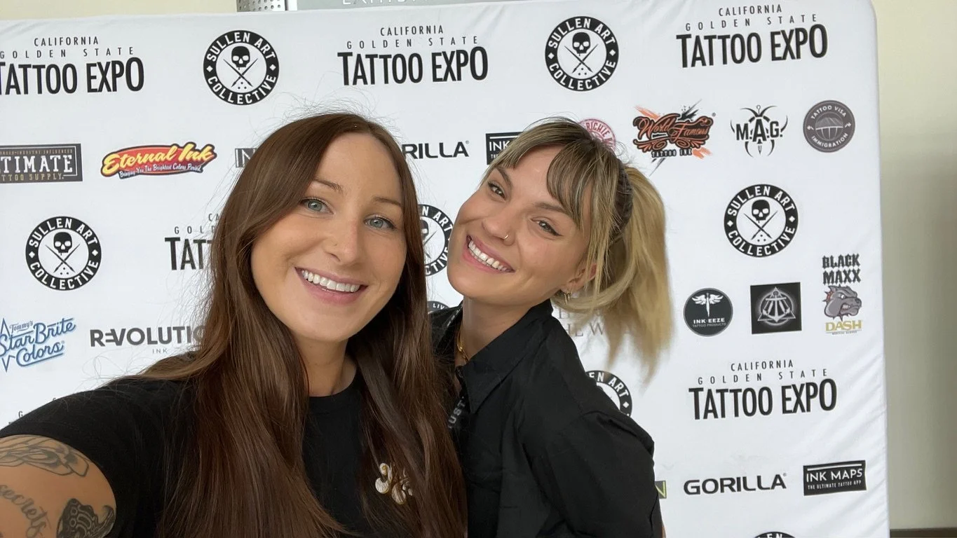 Two women smiling and taking a selfie at the California Golden State Tattoo Expo, with a branded backdrop behind them.
