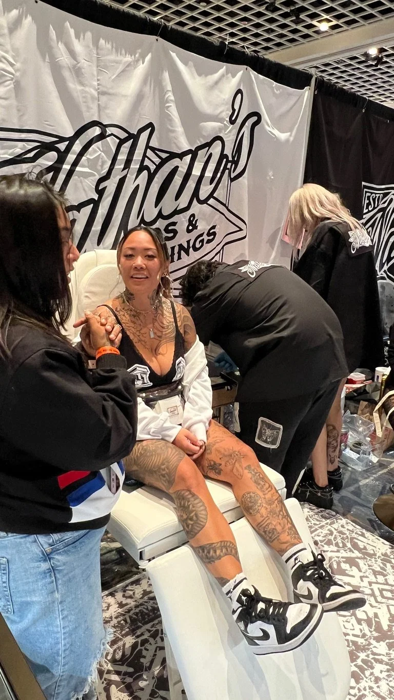 A woman with tattoos on her legs and chest is sitting on a chair at a tattoo convention, surrounded by people and equipment. She appears to be getting a tattoo, and the background includes a large black and white banner with text.